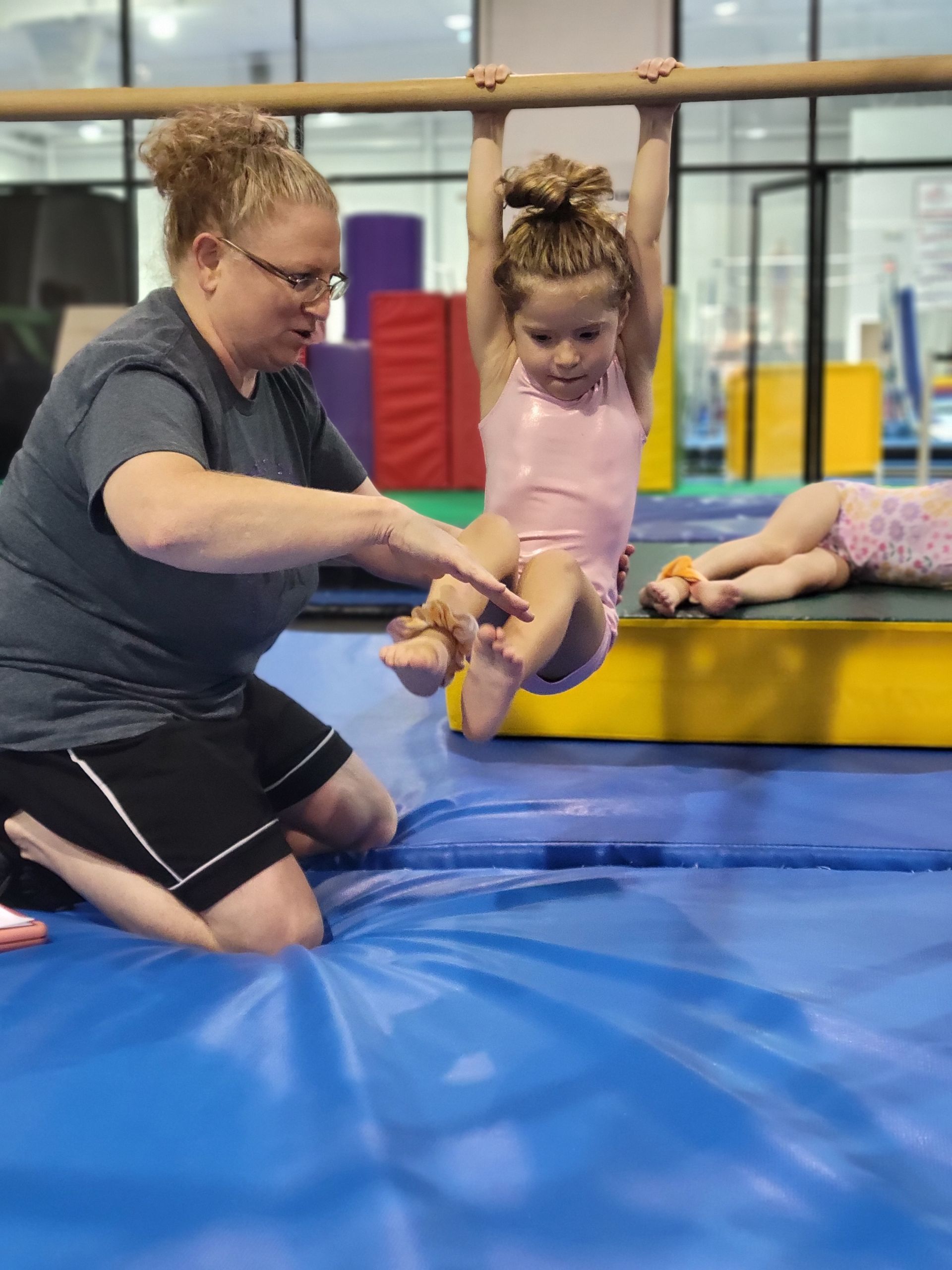 Woman helping a child hang from a bar in a gym. Child in pink leotard, blue mat, and other equipment in the background.