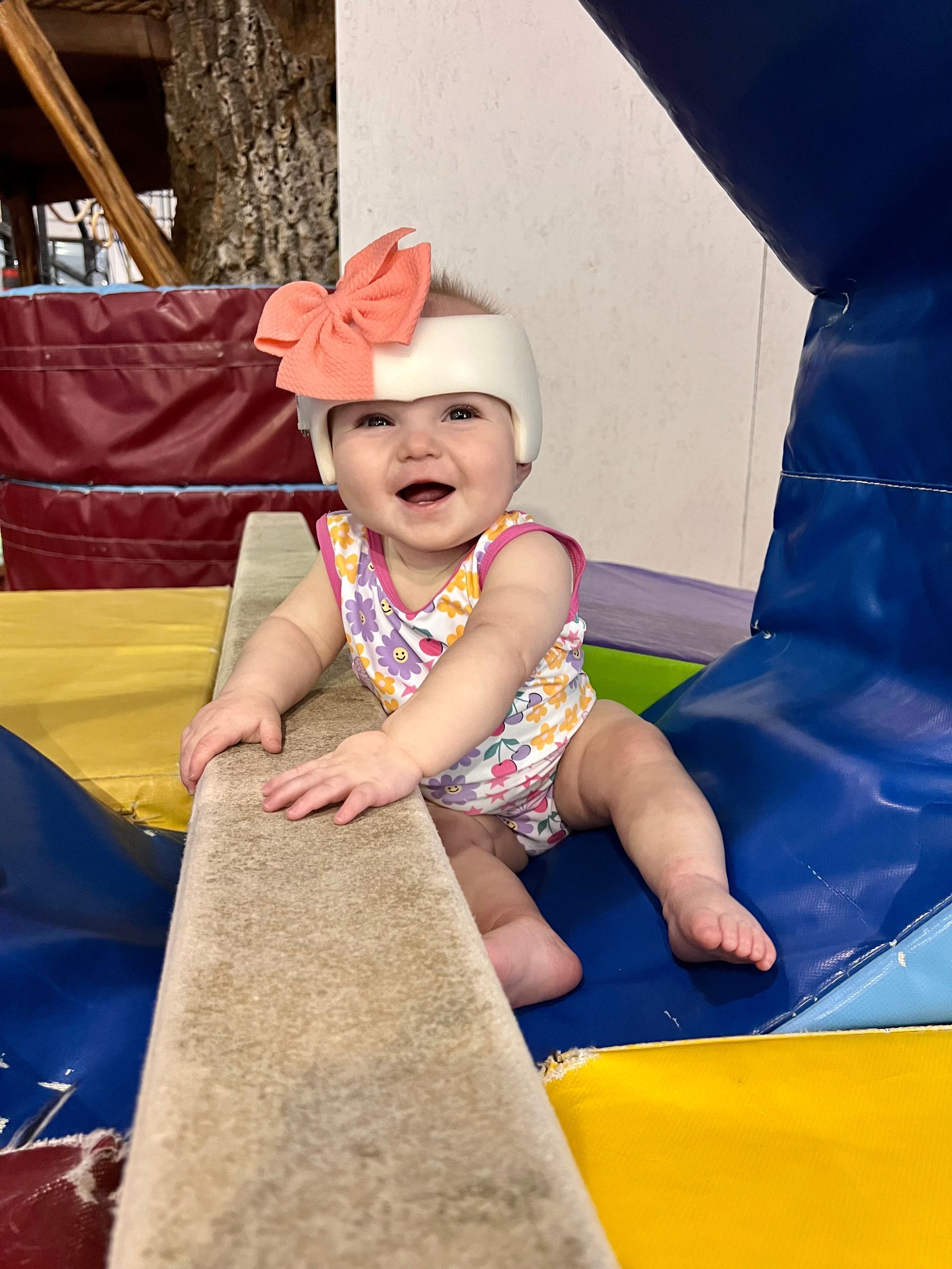 Smiling baby wearing helmet with orange bow on a colorful play structure.