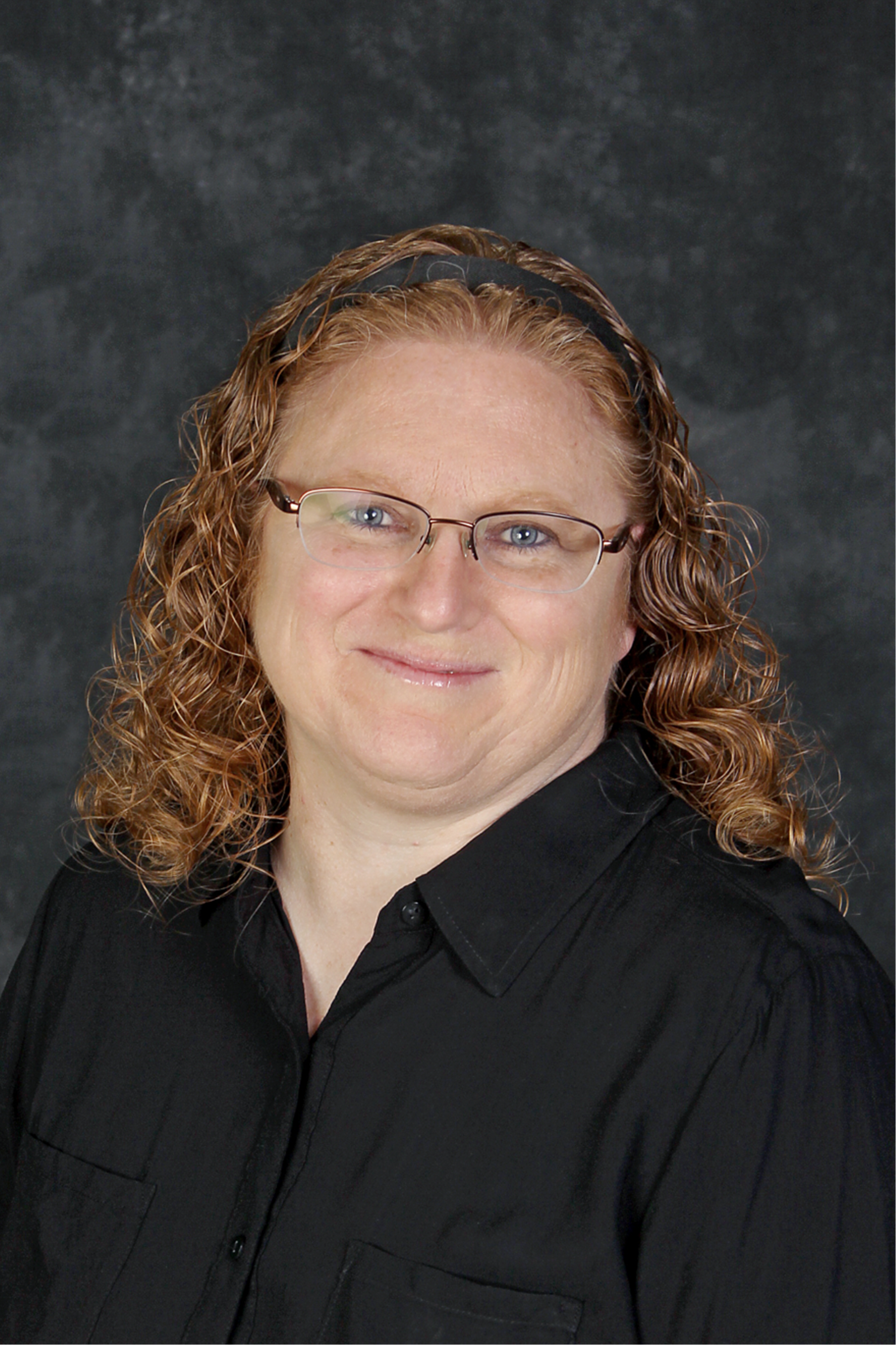 Woman with curly red hair, glasses, wearing a black shirt, smiling, against a mottled gray background.