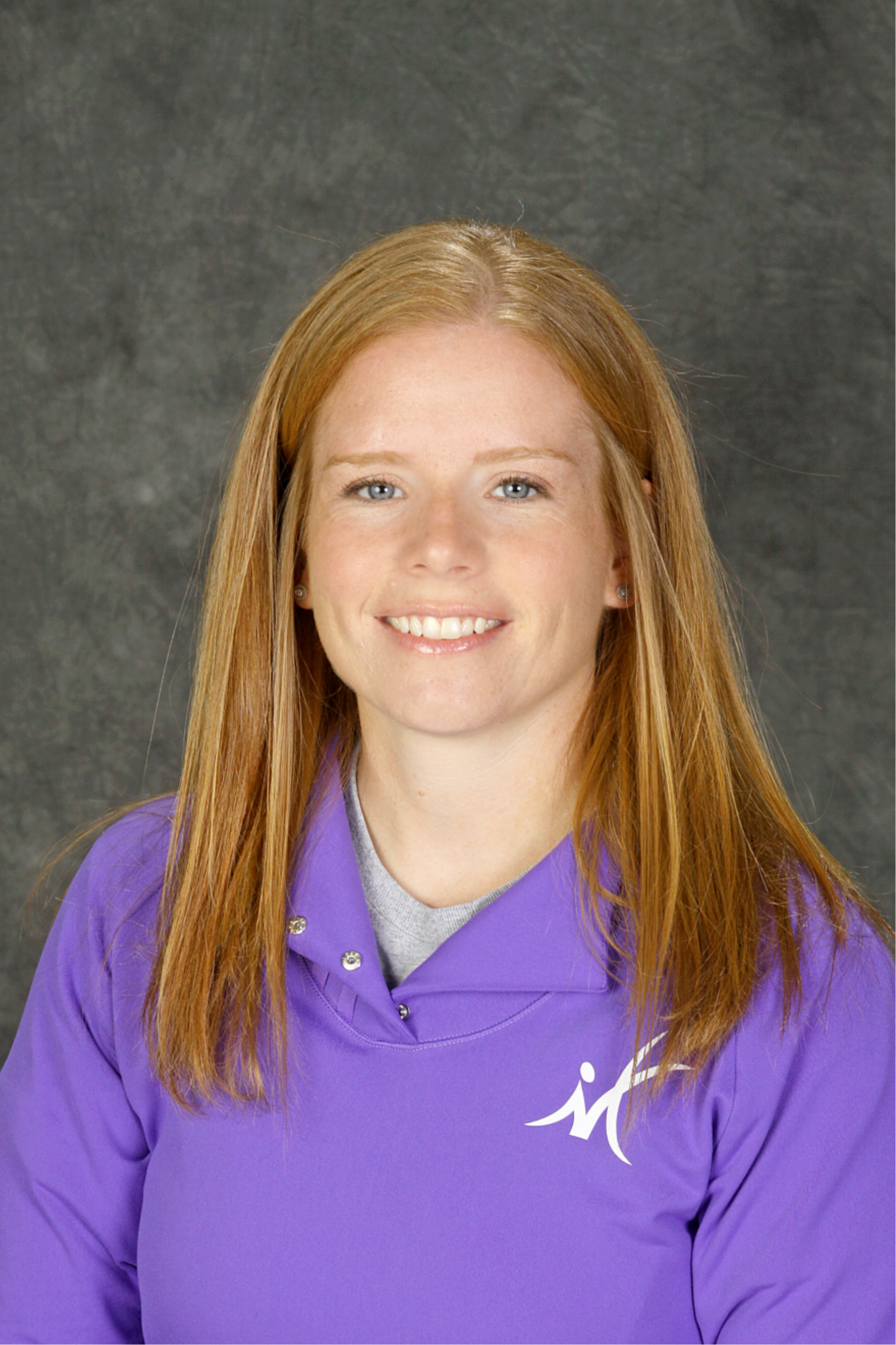 Woman with red hair smiling, wearing a purple jacket with a logo, against a gray background.