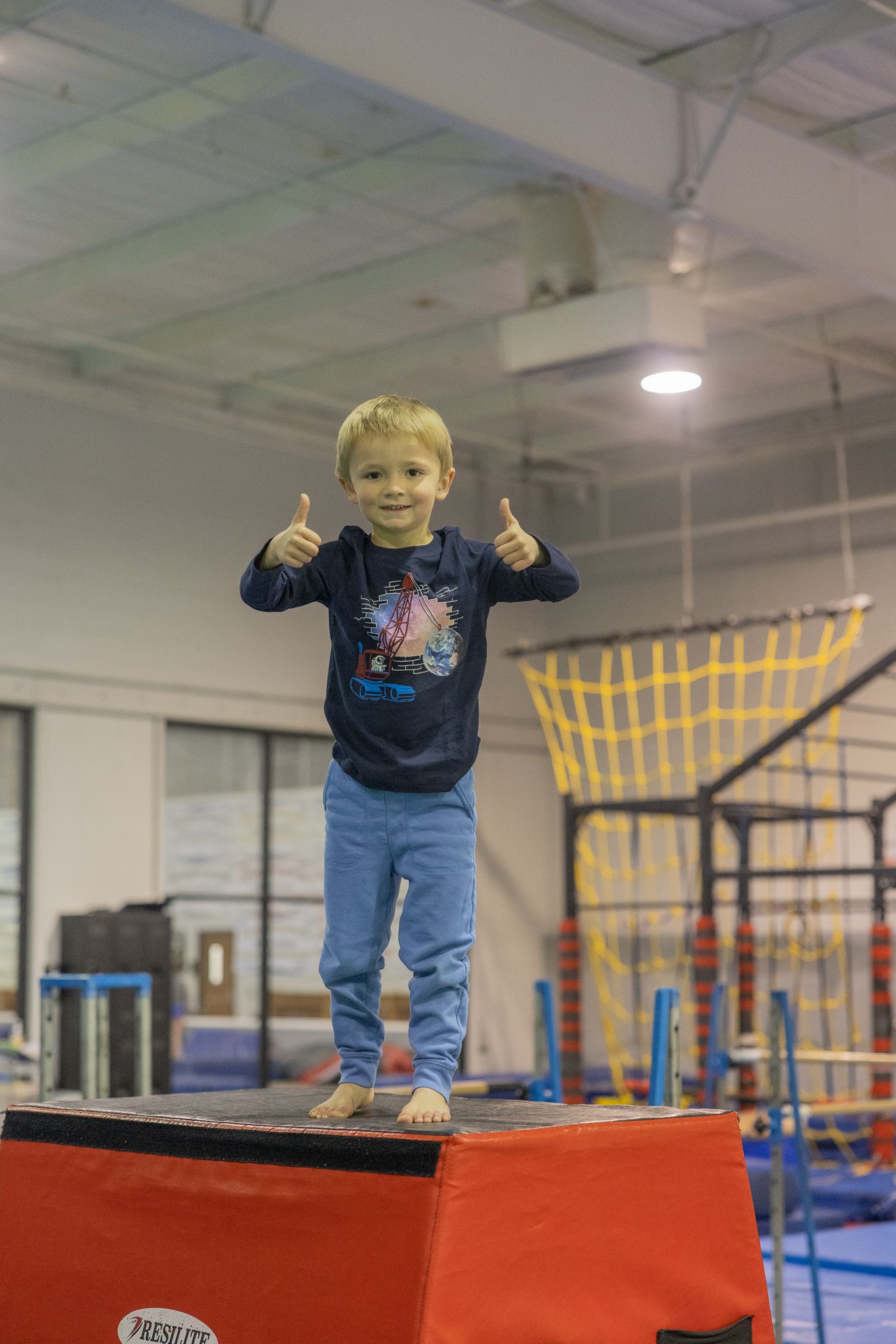 Young boy giving thumbs up, standing on a red box in a gym.