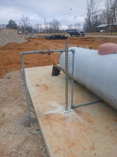 Industrial pipe and tank on a concrete pad at a construction site with dirt and parked trucks in the background