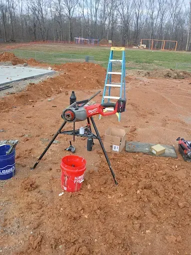 Construction area with a miter saw on a stand, orange bucket, ladder, and chairs in a muddy yard.