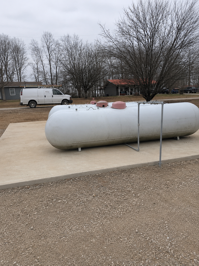 Large gray propane tank on a gravel lot with a white van and bare trees in the background