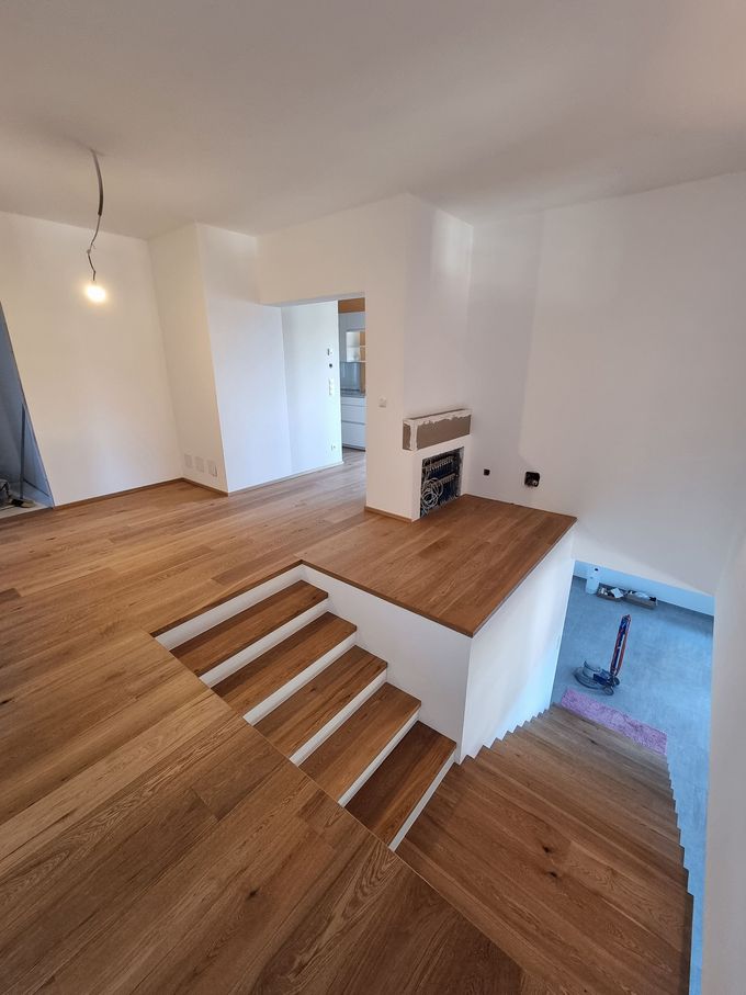 Holzboden mit einer kleinen Treppe, die zu einer erhöhten Plattform mit Kamin führt; in der Ferne ist ein Türrahmen sichtbar.