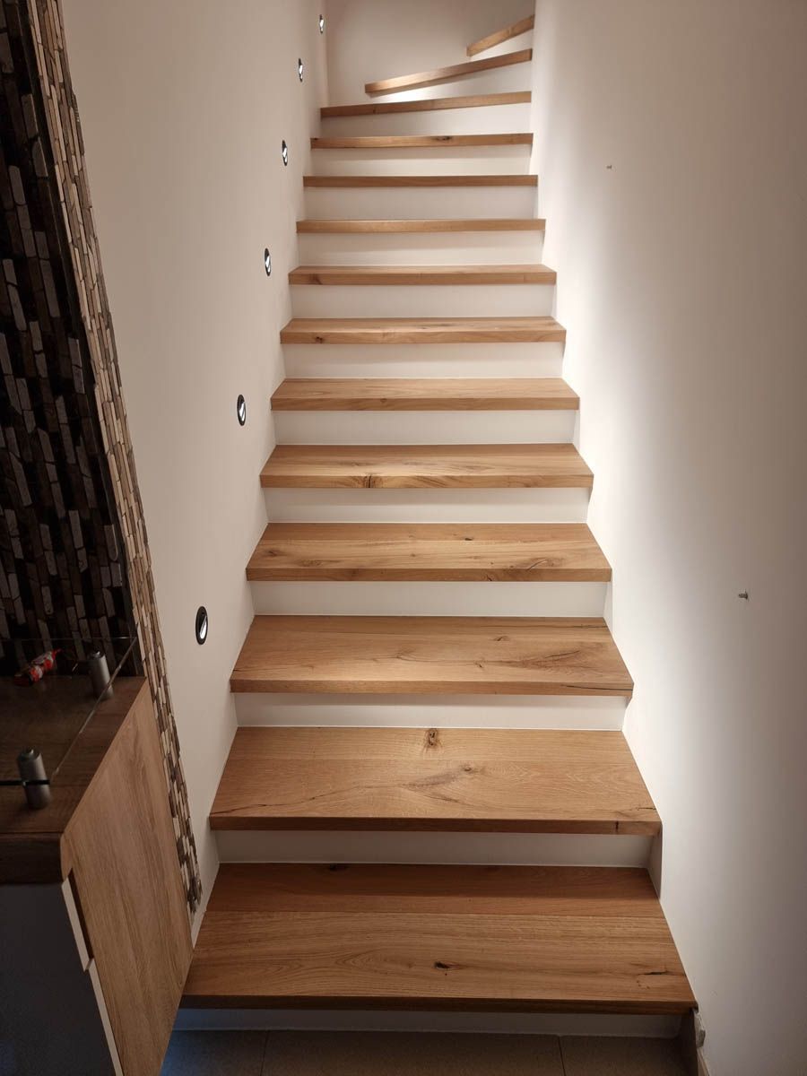 Zwischen weißen Wänden nach oben führende Holztreppe mit weißen Setzstufen und Einbauleuchten.