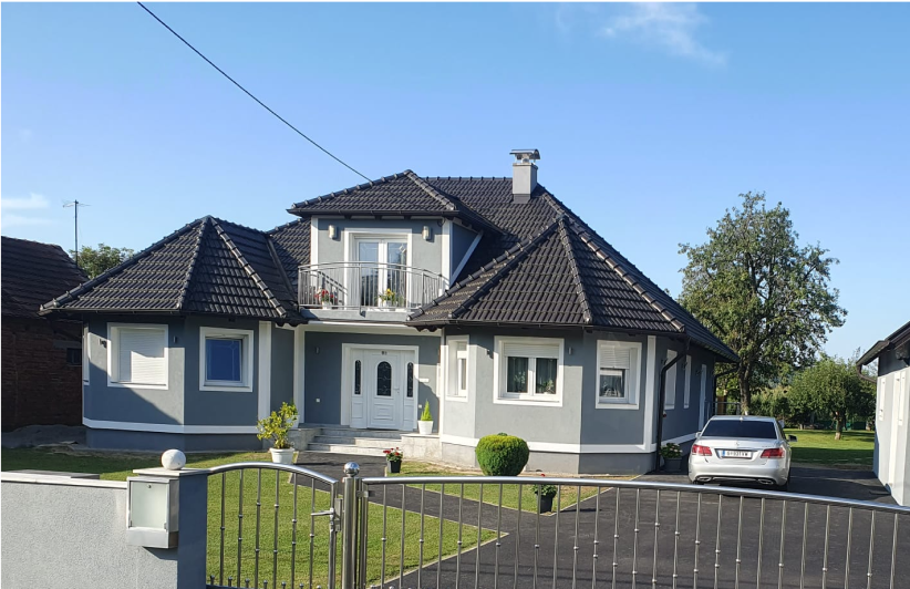 Graues Haus mit dunklem Dach, weißen Zierleisten, kleinem Balkon, davor geparktes Auto.