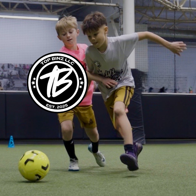 Two male players playing indoor soccer, chasing a yellow ball on turf. Top Binz Logo on top of image