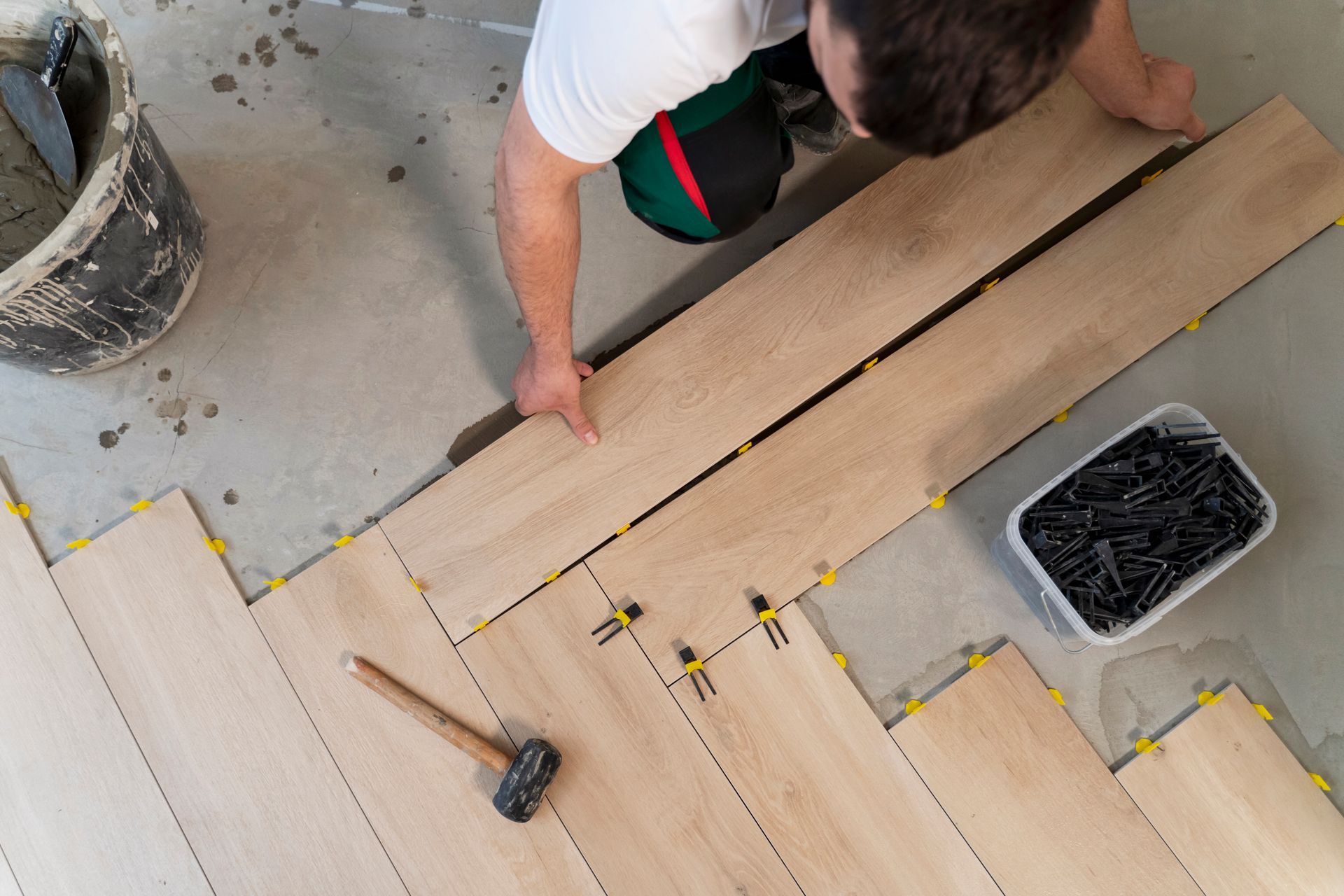 Person kneeling on a floor installing light wood planks with a hammer and nails nearby.