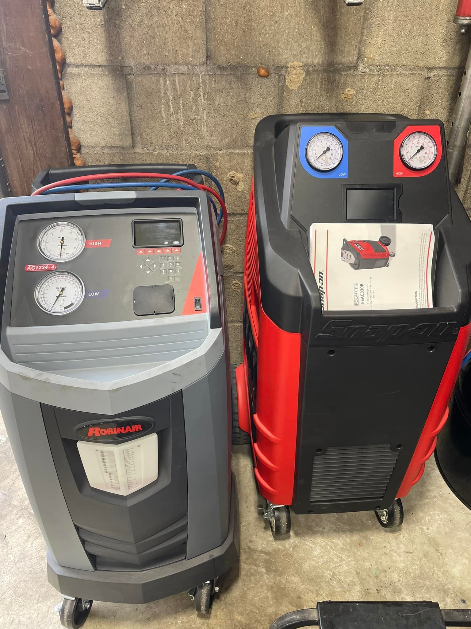 Two air compressors, one gray and one black-red, standing against a brick wall outdoors.