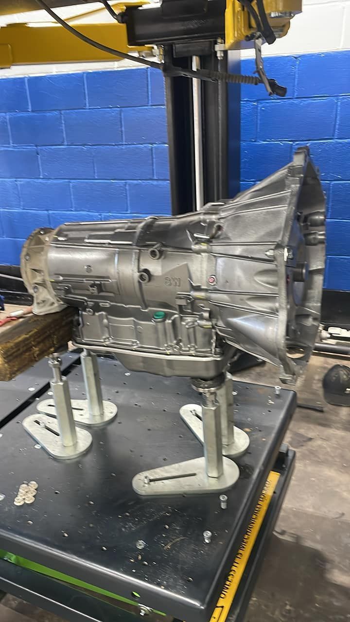 Silver transmission mounted on a stand in a workshop, with a blue brick wall in the background.