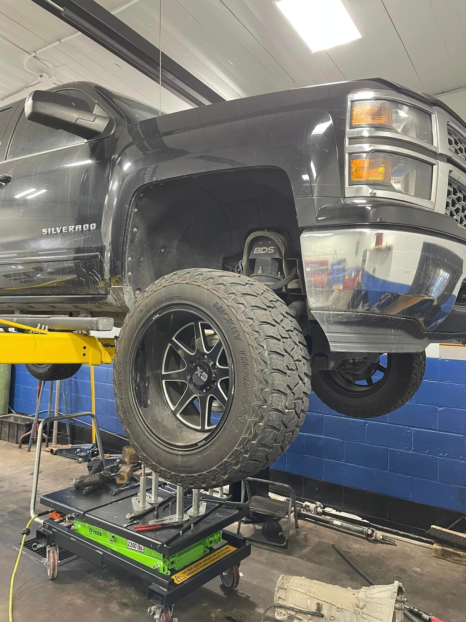 Black pickup truck raised on a shop lift in a garage, front wheel removed during maintenance.