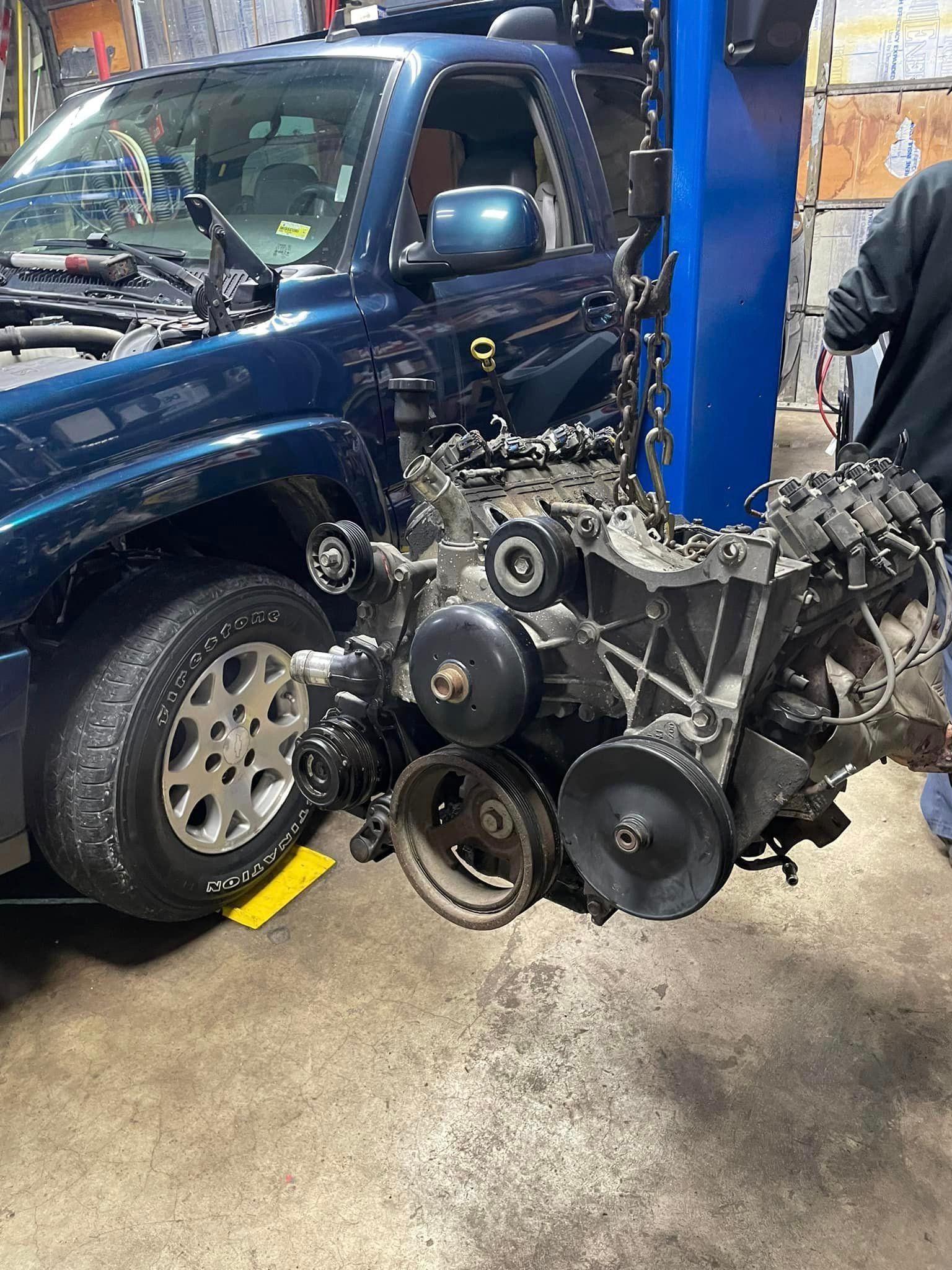 Blue SUV in a garage with its engine removed and sitting on the floor beside it.