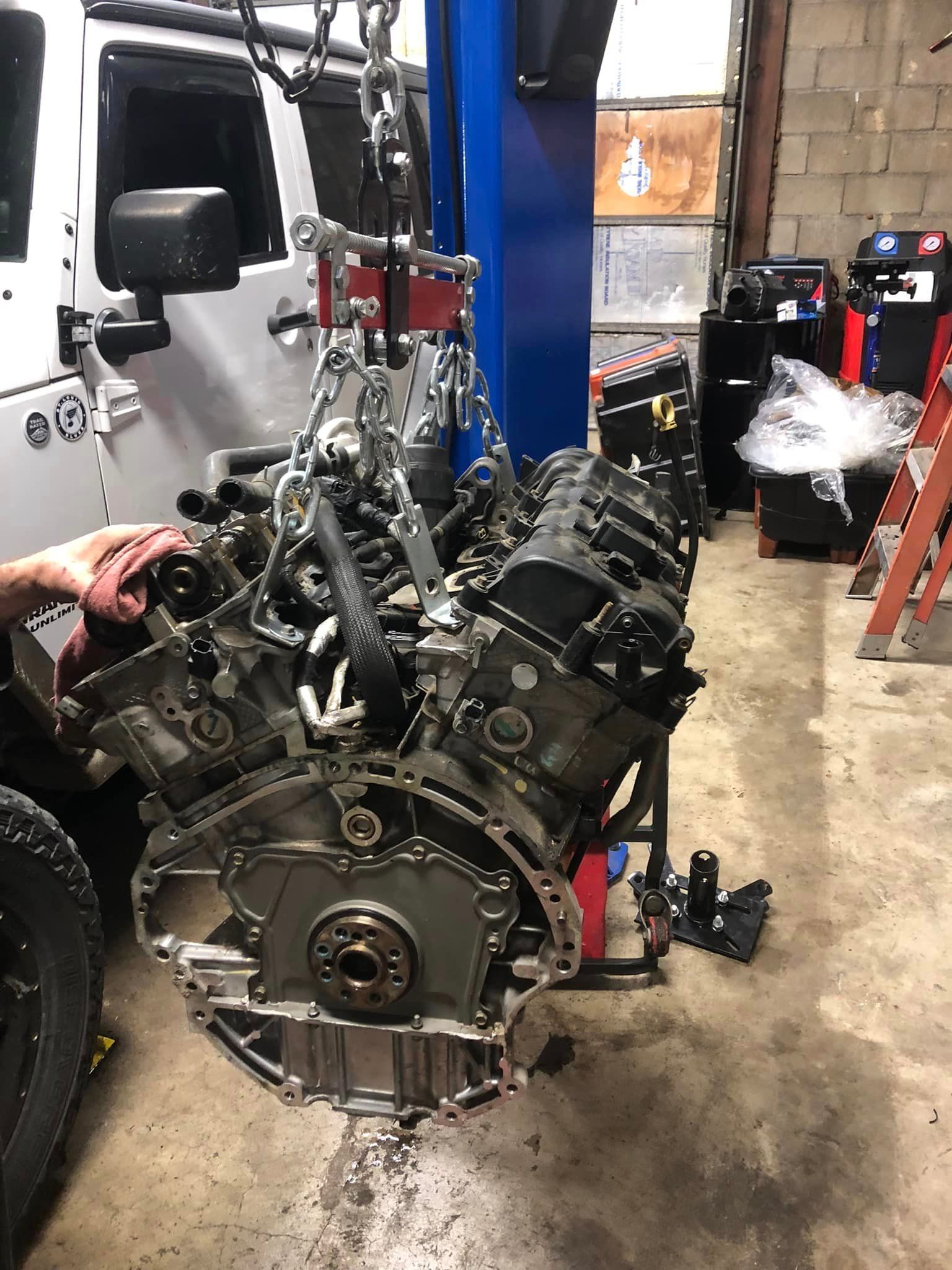 Car engine on a hoist in a workshop, with mechanic’s hand visible on the left.
