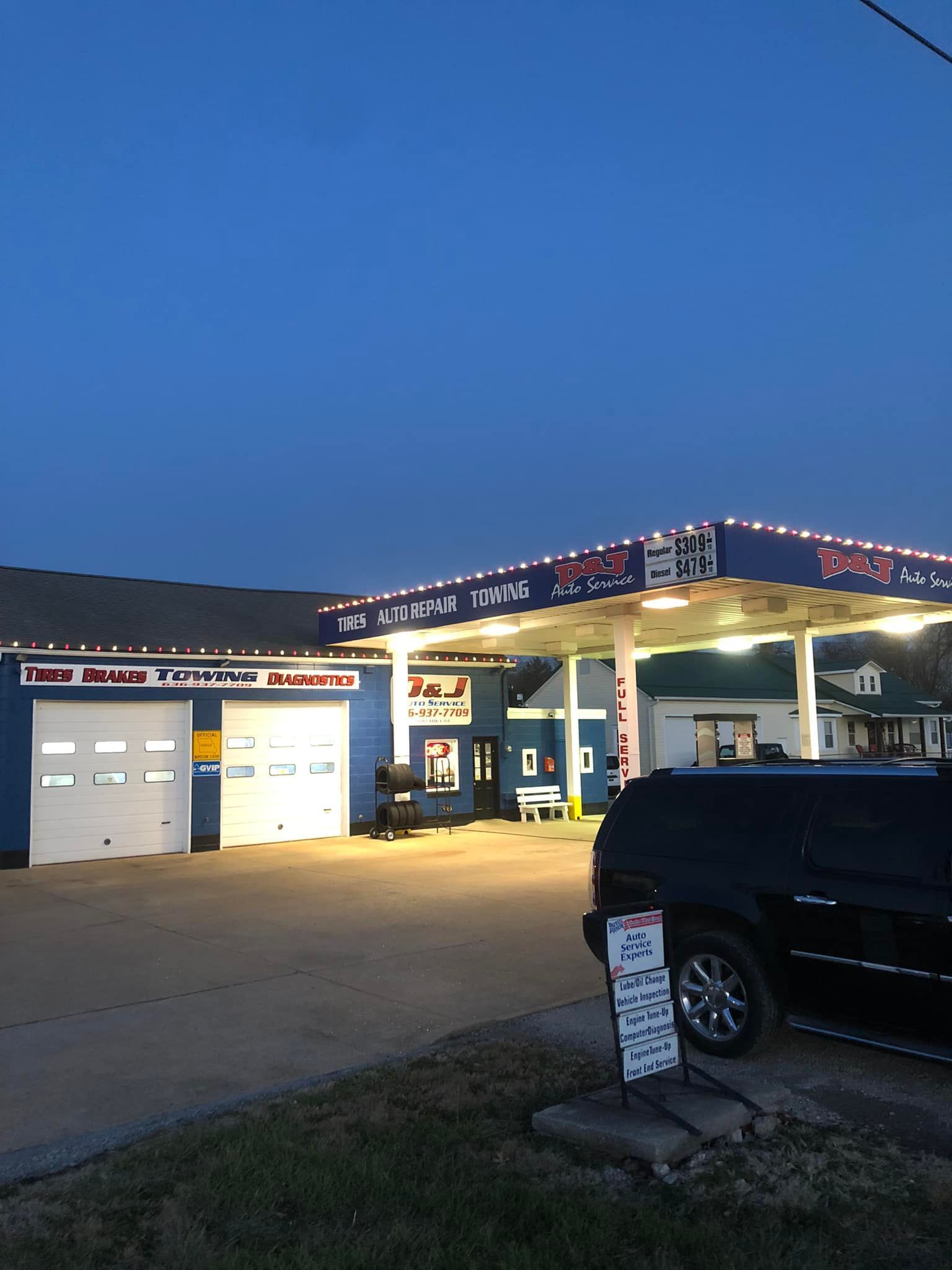 Small auto repair shop at dusk with lit signs, open garage doors, and a black SUV parked outside.