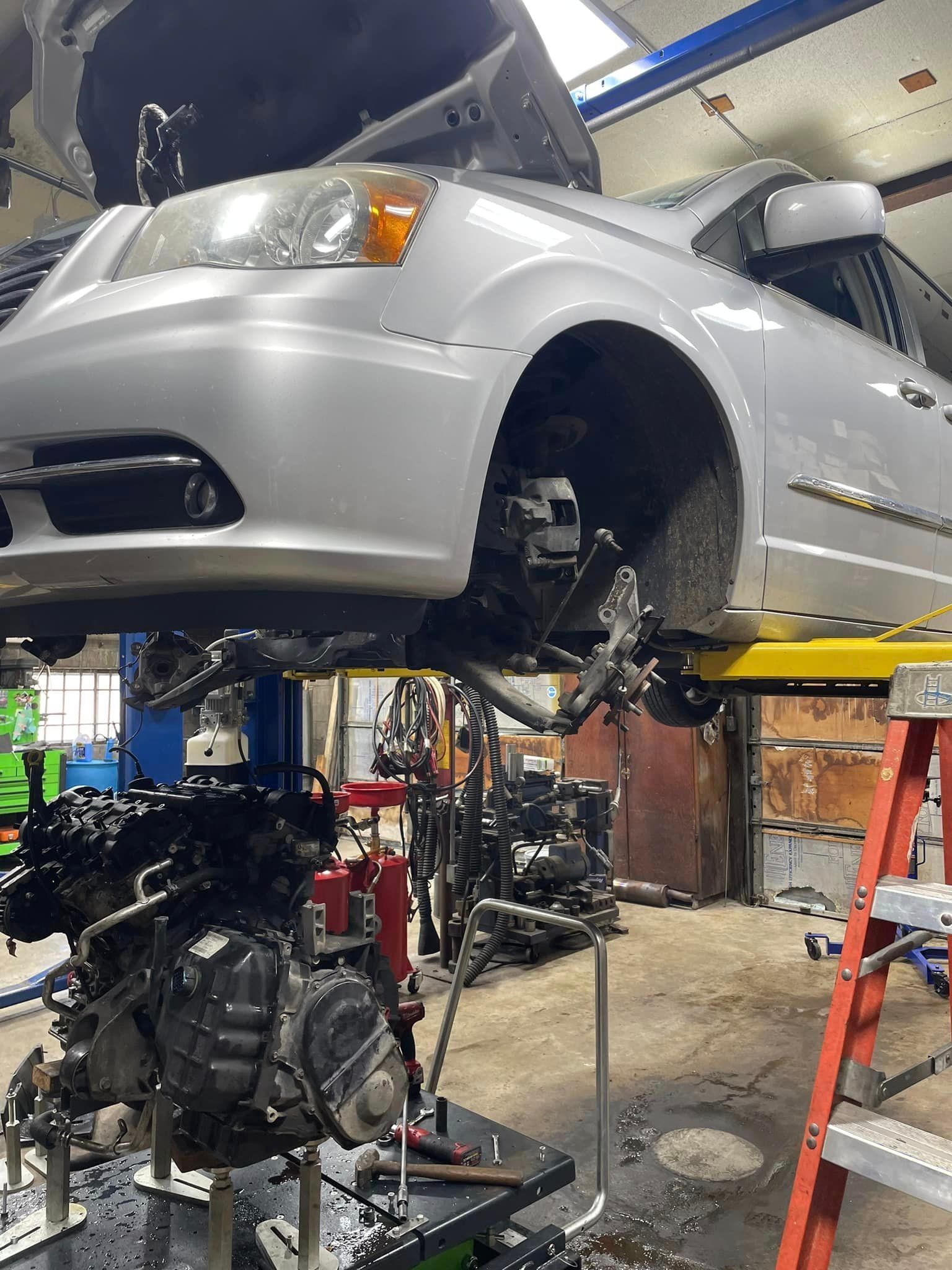 Silver car raised in auto shop, front wheel removed, with engine and repair tools below.