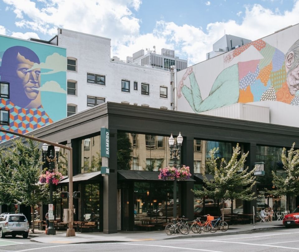 Street scene with modern building featuring large windows, surrounded by trees, parked bikes, and colorful murals on adjacent buildings.