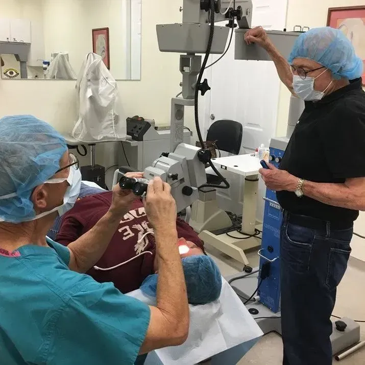 Two people in surgical attire performing an eye exam. One looks through binoculars. Another adjusts equipment.