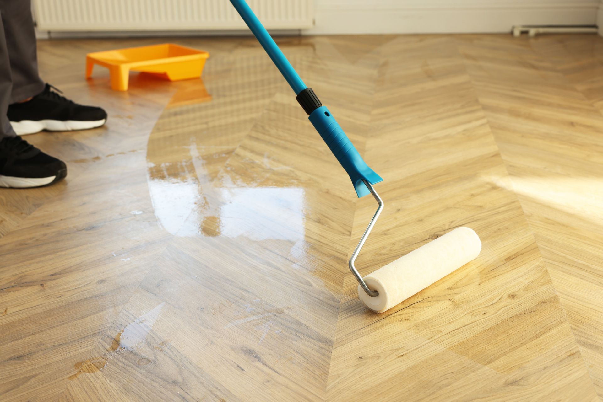 Person applying a glossy sealant to a light wood parquet floor with a roller.