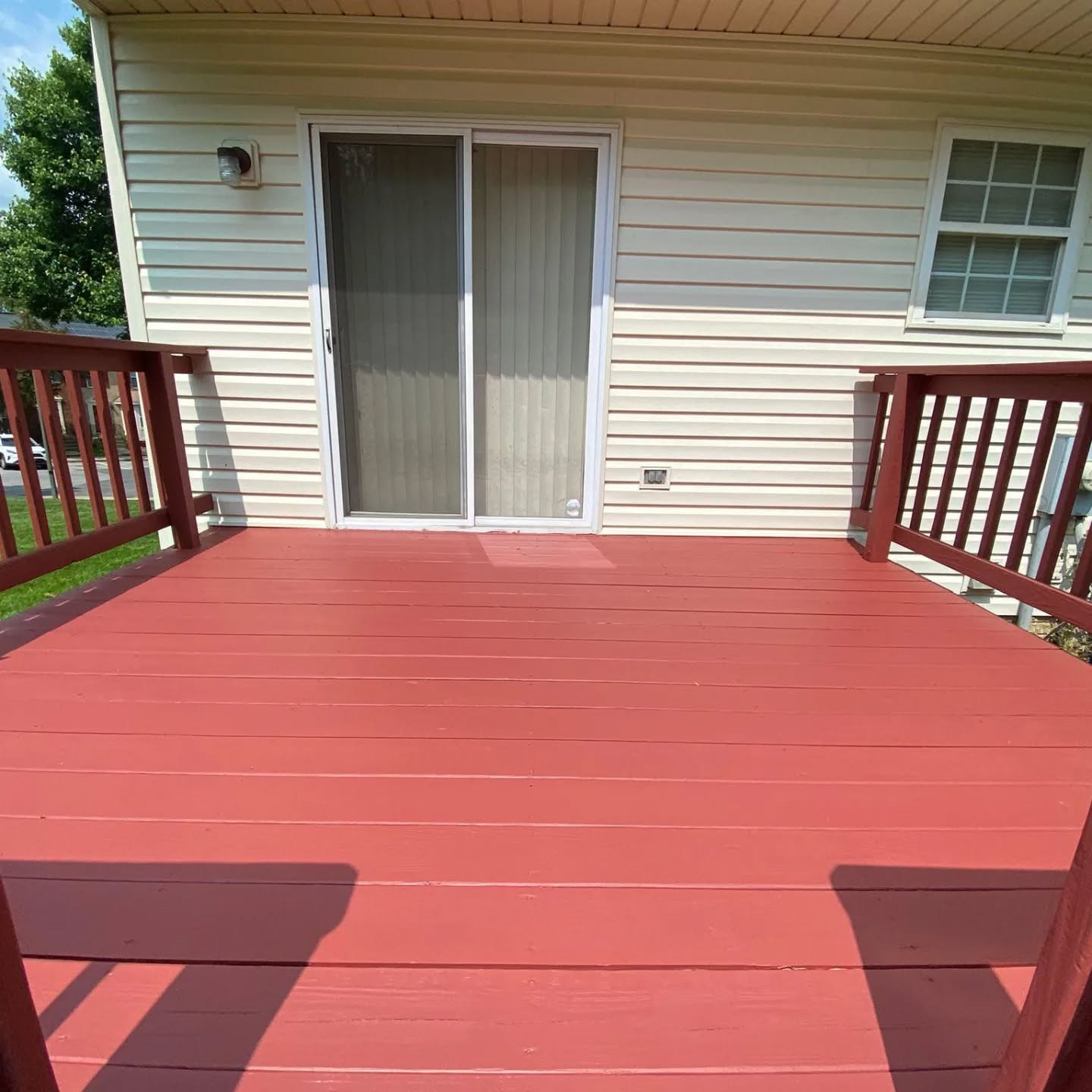 Red painted wooden deck with railing, sliding glass door, and a white house with a window.