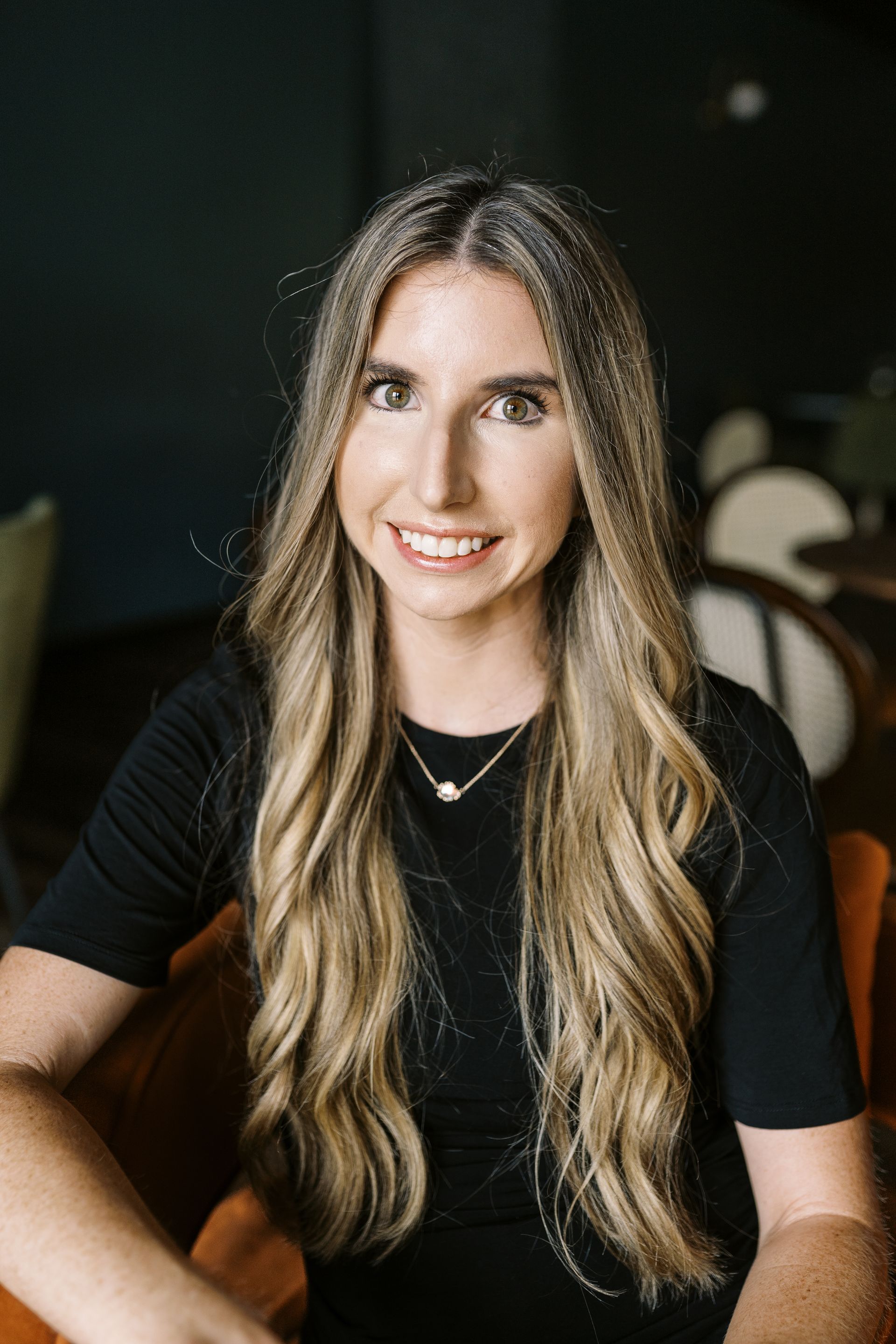 A person with long blonde hair and a necklace smiling, sitting in a dark, softly lit room with chairs in the background.