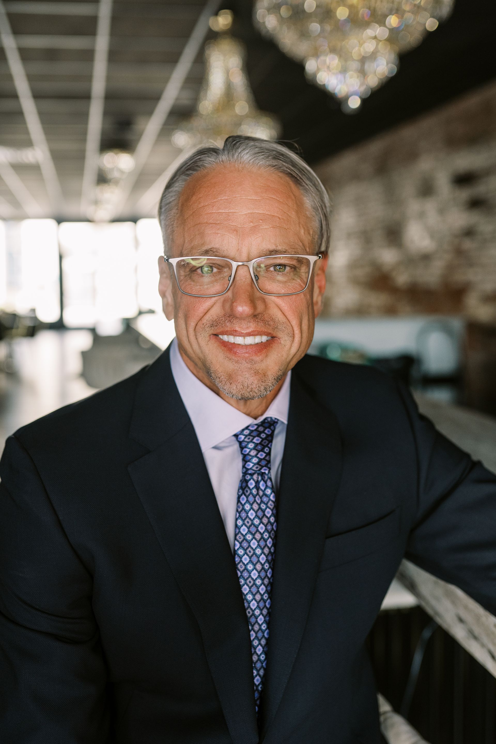 A smiling person with glasses wearing a dark suit, light shirt, and patterned tie, posing in a modern interior.