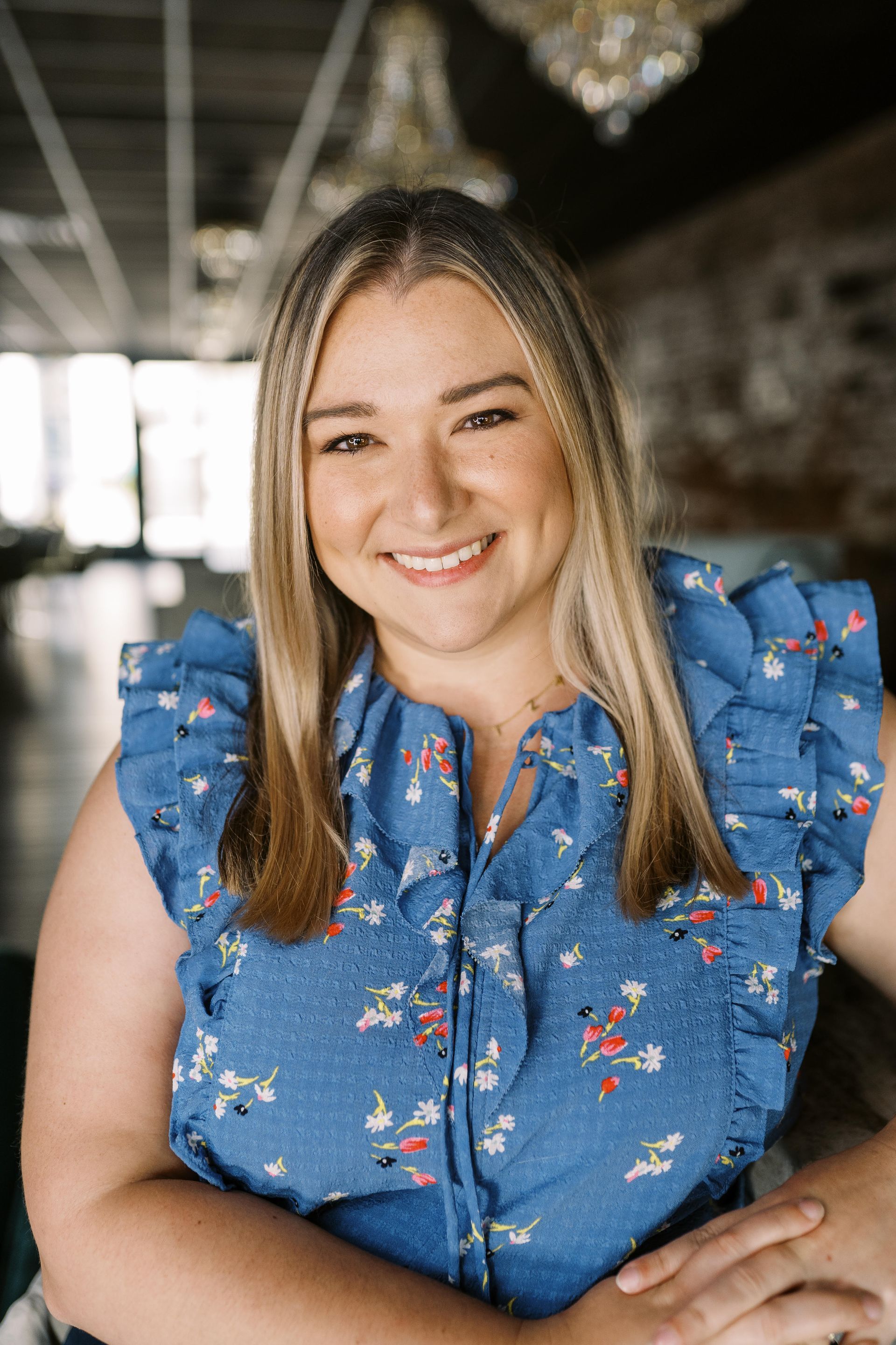 A smiling person with long hair wearing a blue ruffled floral top, standing in a brightly lit indoor space.