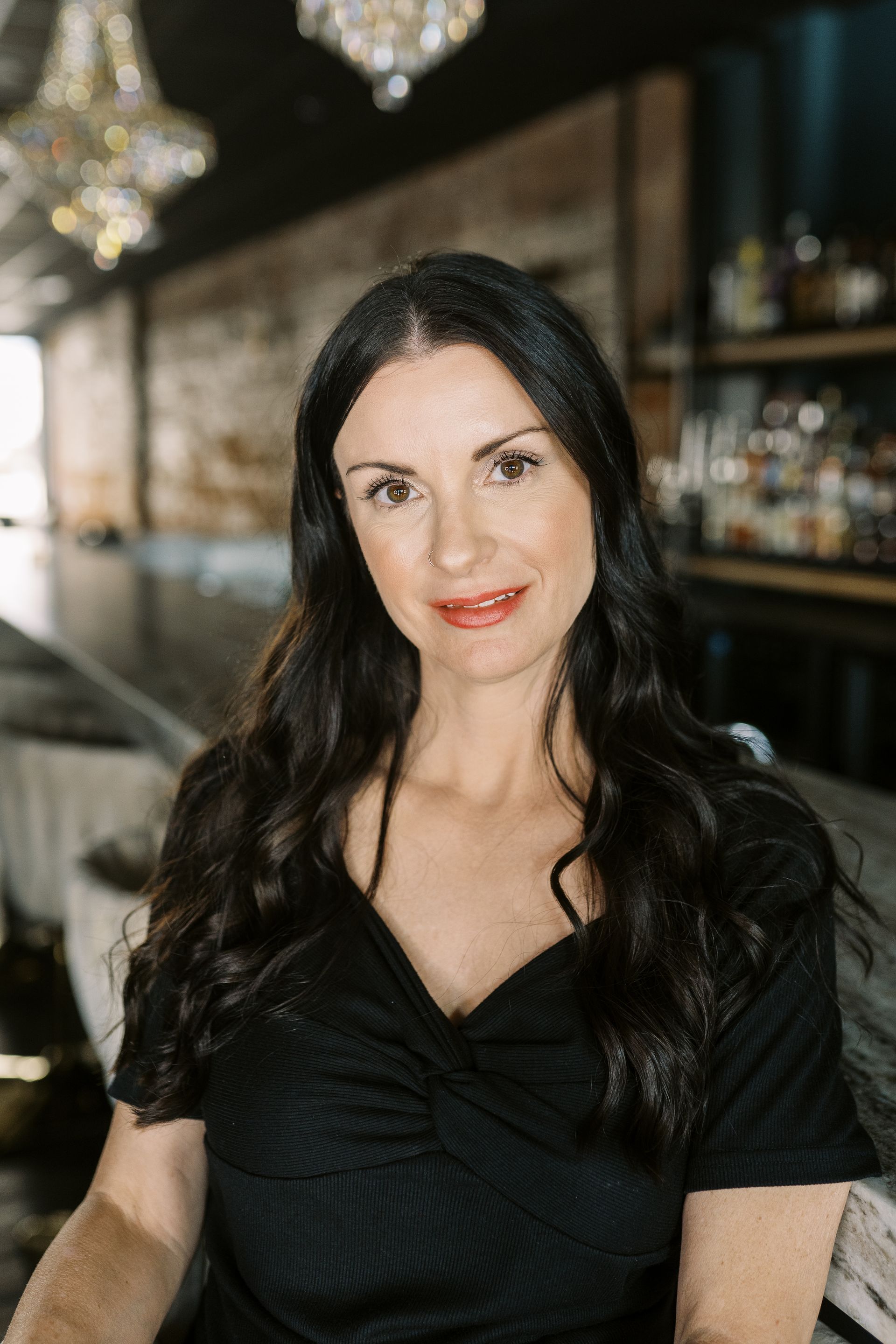 A person with dark hair and a neutral expression in a black top, seated in a dimly lit bar with crystal chandeliers.