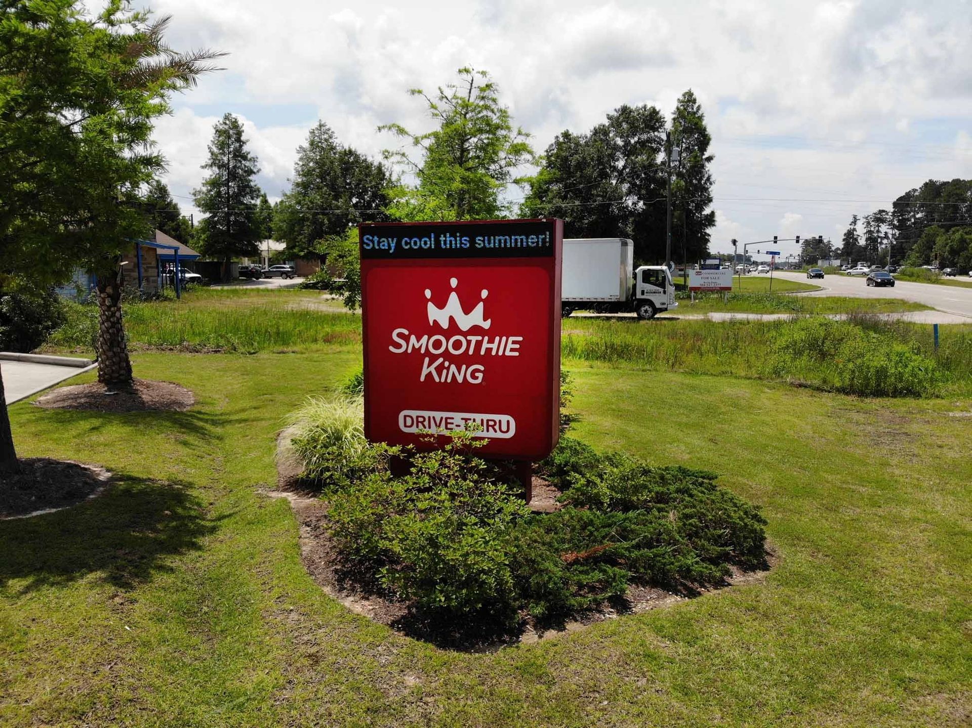 Smoothie King sign in a grassy area, red and white logo, drive-thru, trees and a truck in the background.