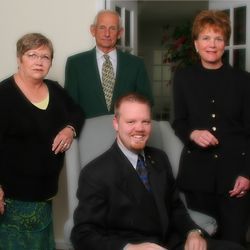 Four people posing indoors, three standing and one seated, all dressed in dark formal clothing.
