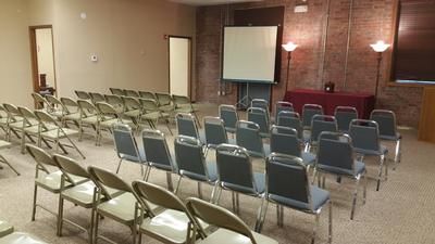 Empty conference room with rows of folding chairs facing a projector screen and lectern on a brick wall stage