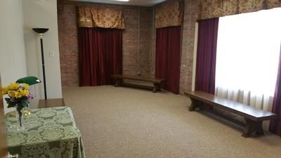 Empty carpeted room with burgundy curtains, long wooden benches, and a small flower arrangement on the left.