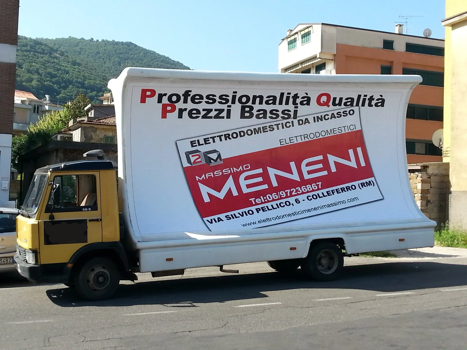Un camion pubblicitario giallo parcheggiato in strada, con un grande cartello curvo che pubblicizza gli elettrodomestici Meneni.