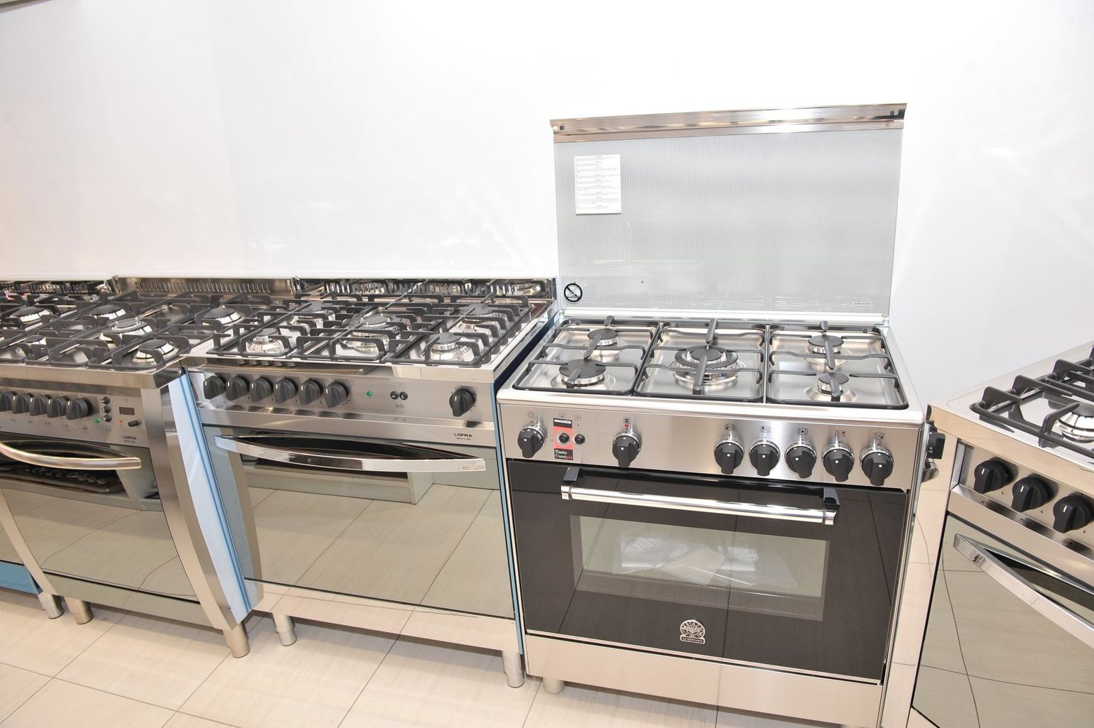 In uno showroom sono disposte in fila diverse cucine a gas professionali in acciaio inox con bruciatori e forni.