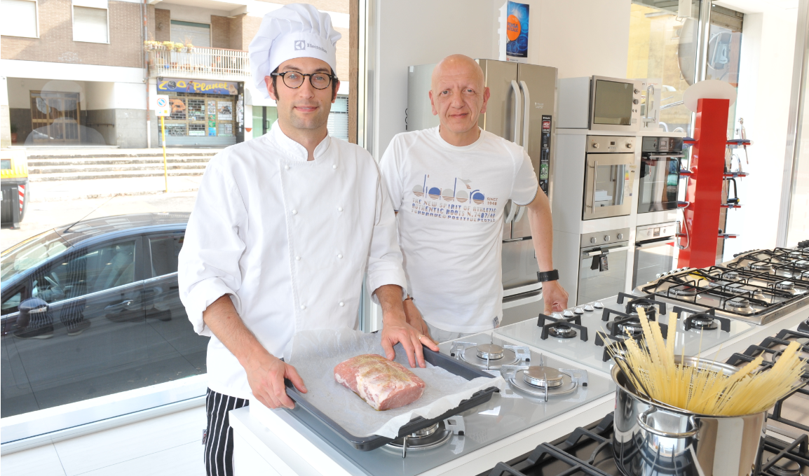 In una sala espositiva, due uomini sono in piedi dietro un bancone da cucina: uno indossa una divisa da chef e tiene in mano una teglia con della carne.