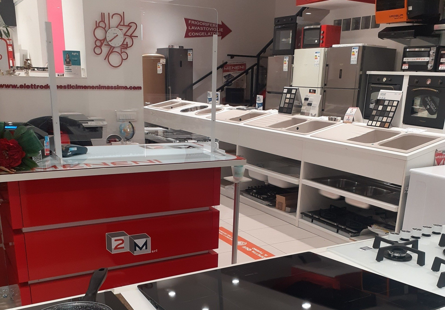 Uno showroom di elettrodomestici con mobili rossi, diversi piani cottura, forni e un grande orologio da parete, il tutto in uno spazio luminoso e moderno.