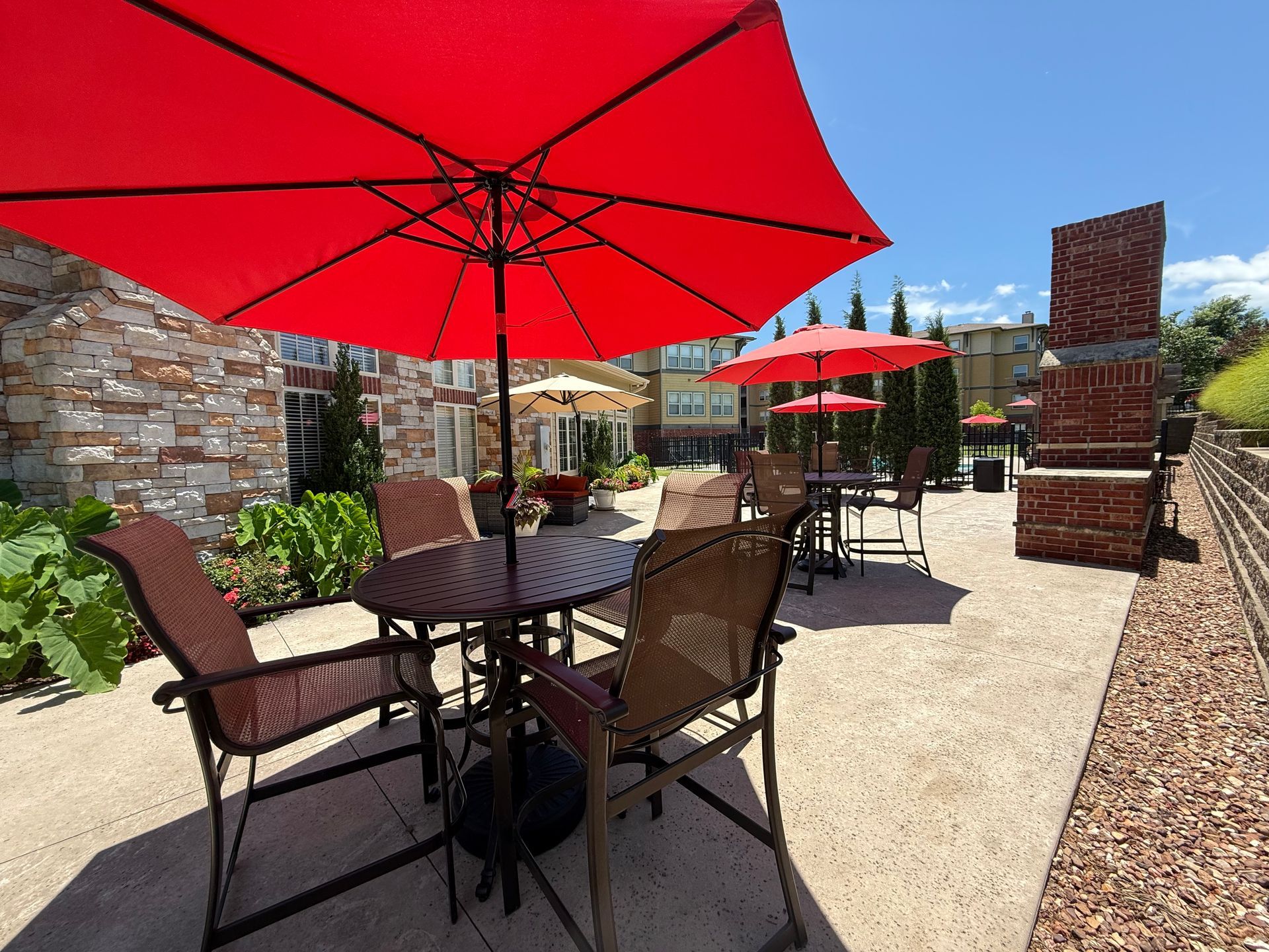 A sunny outdoor patio with metal tables, chairs, and large red umbrellas against a stone building and brick fireplace.