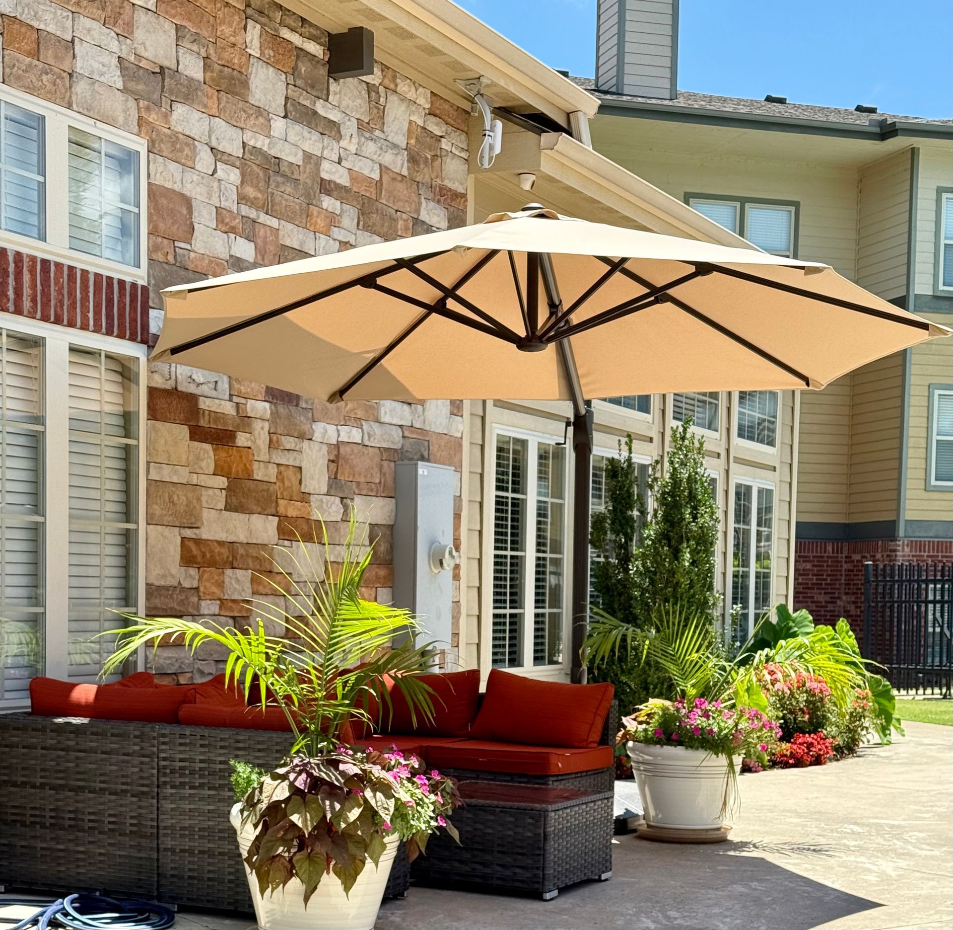 A tan patio umbrella shades a wicker sectional with red cushions and potted plants on a stone patio outside a brick home.