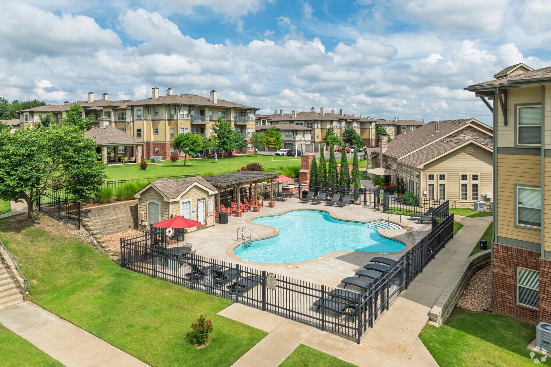 An aerial view of an apartment complex pool area with beige buildings, green lawns, lounge chairs, and a red umbrella.