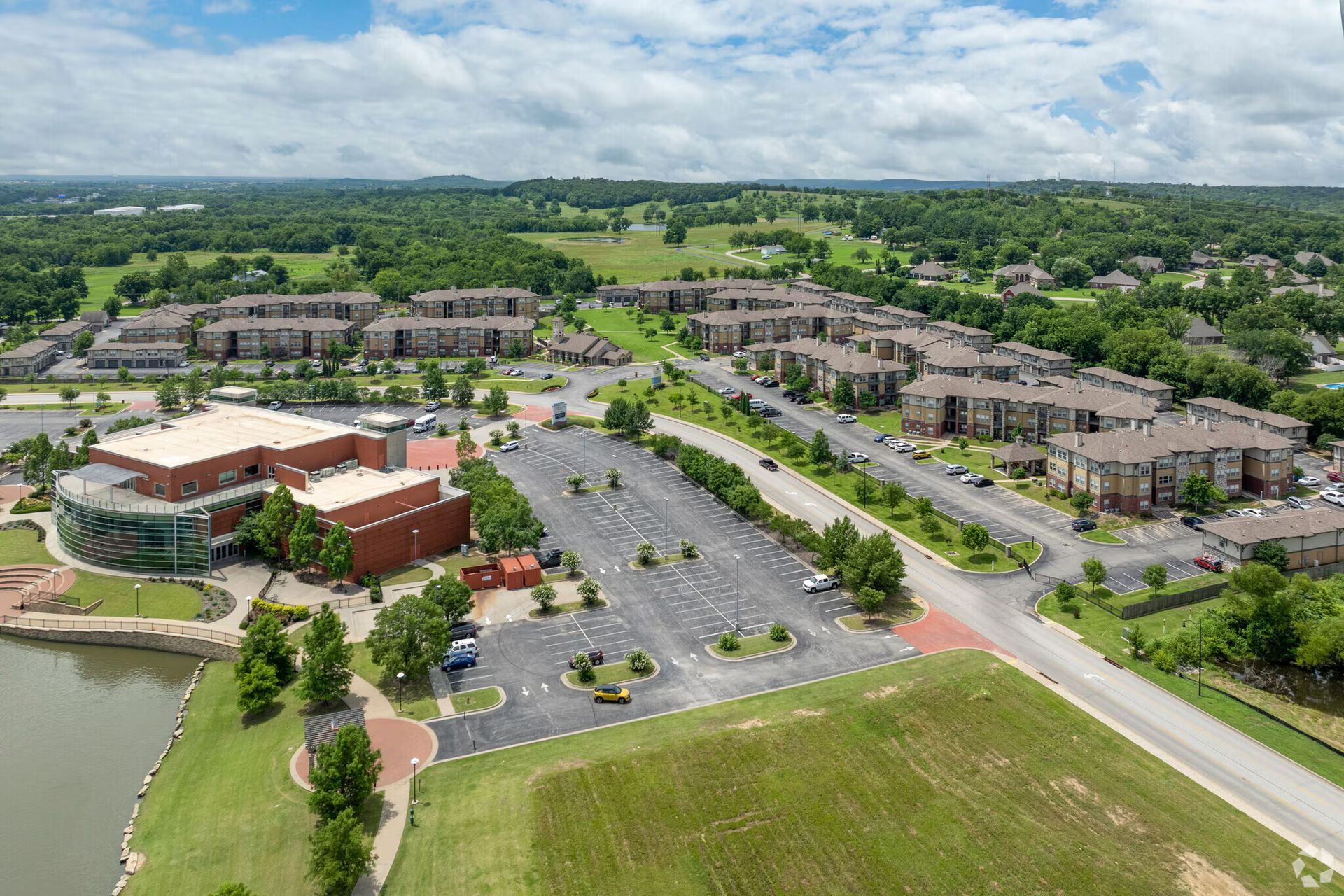 Aerial view of a brown brick building, a parking lot, and adjacent residential apartment complexes near a pond.