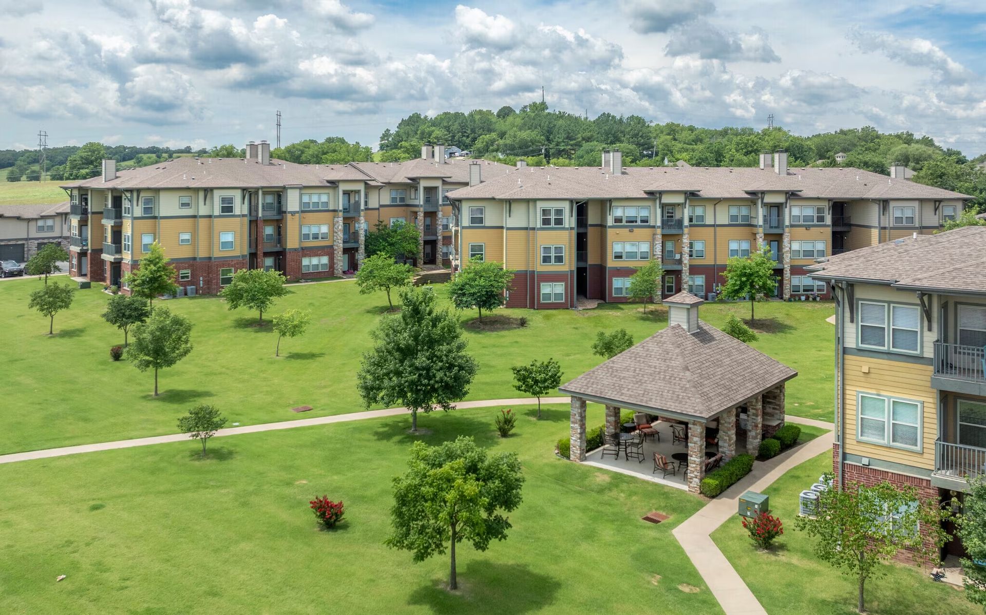 A multi-story apartment complex with a large grassy lawn, trees, and a stone gazebo on a sunny, cloudy day.