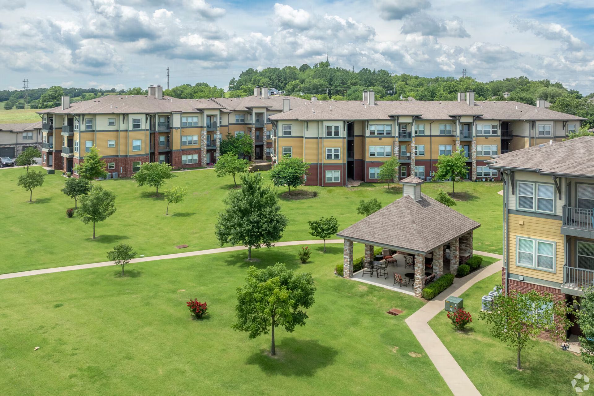 Multistory apartment complex with tan and brick siding, surrounding a green lawn with a gazebo and paved walkways.