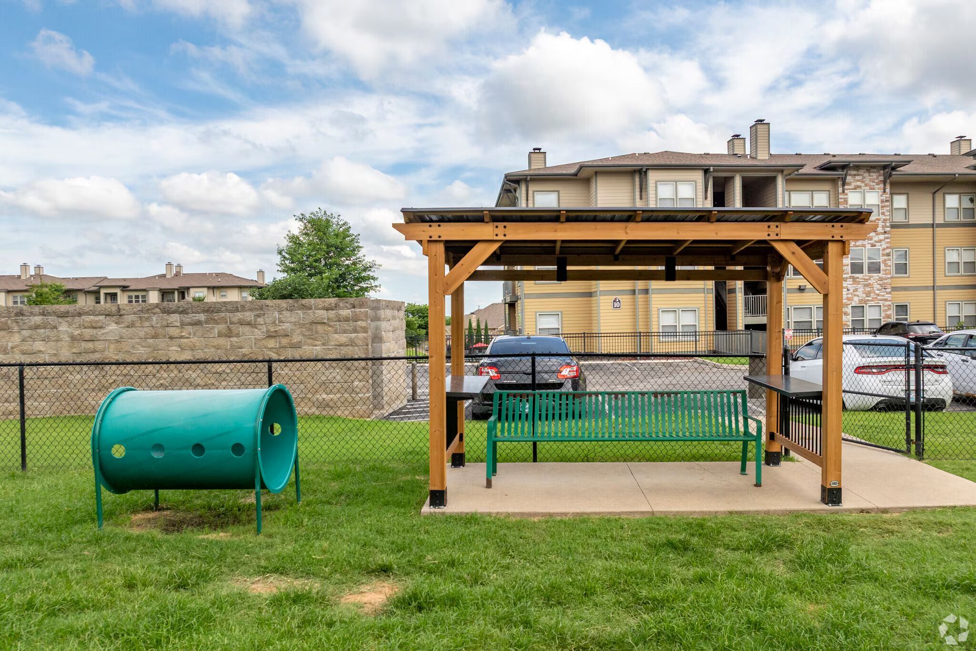 A dog park features a green agility tunnel, a green bench under a wooden pergola, and apartment buildings in the back.