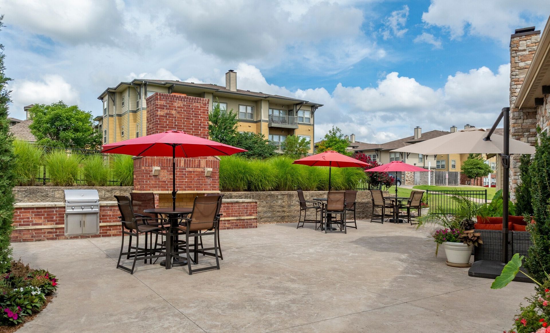 Outdoor patio area with dining tables, red and beige umbrellas, a stone fireplace, grill, and landscaped surroundings.