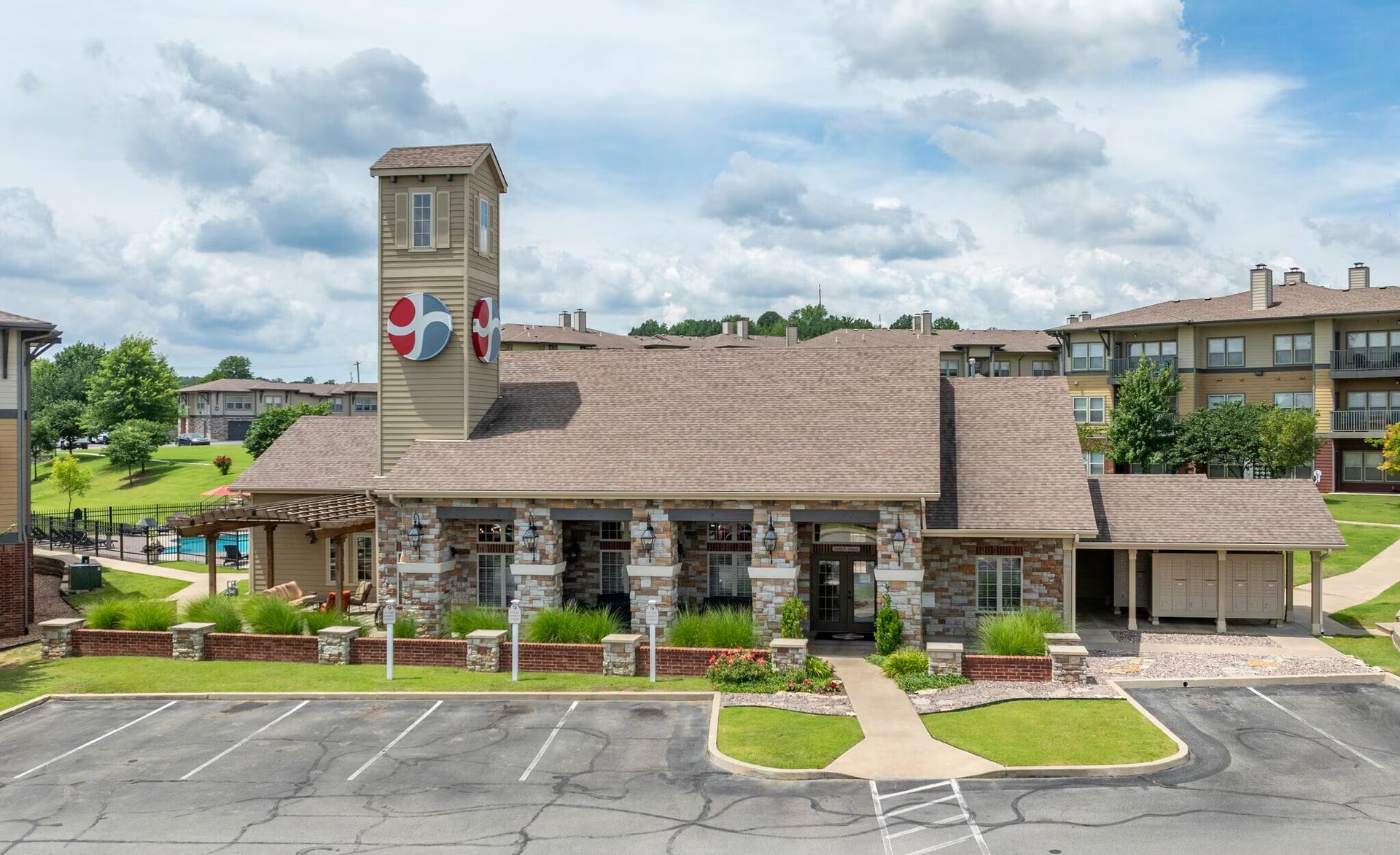 A multi-story beige stone clubhouse with a prominent tower, landscaping, and a front parking lot under a cloudy sky.