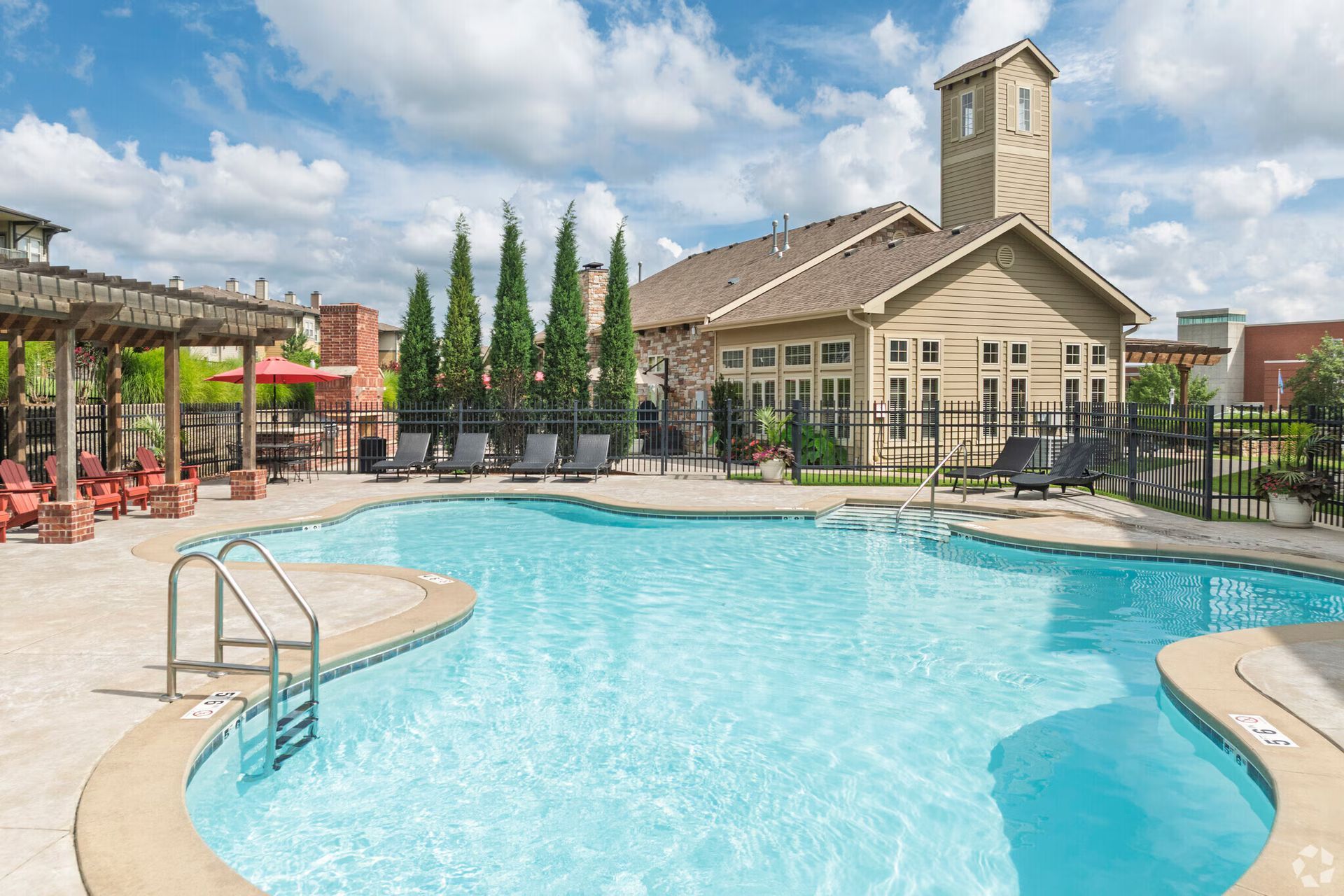 A pool area with a turquoise swimming pool, lounge chairs, a wooden pergola, and a clubhouse building under blue skies.