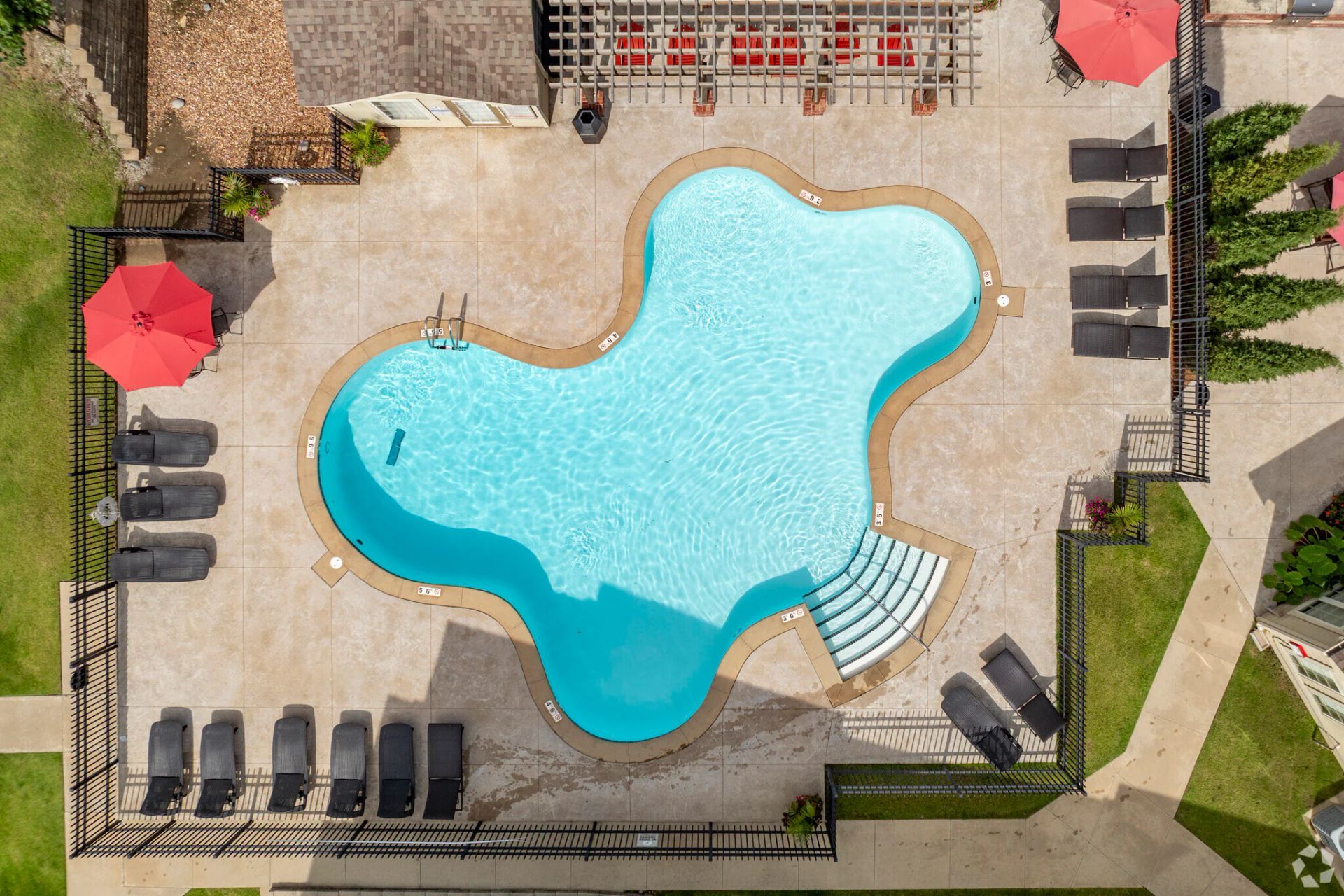 Aerial view of a turquoise swimming pool with lounge chairs and red umbrellas on a stone deck.