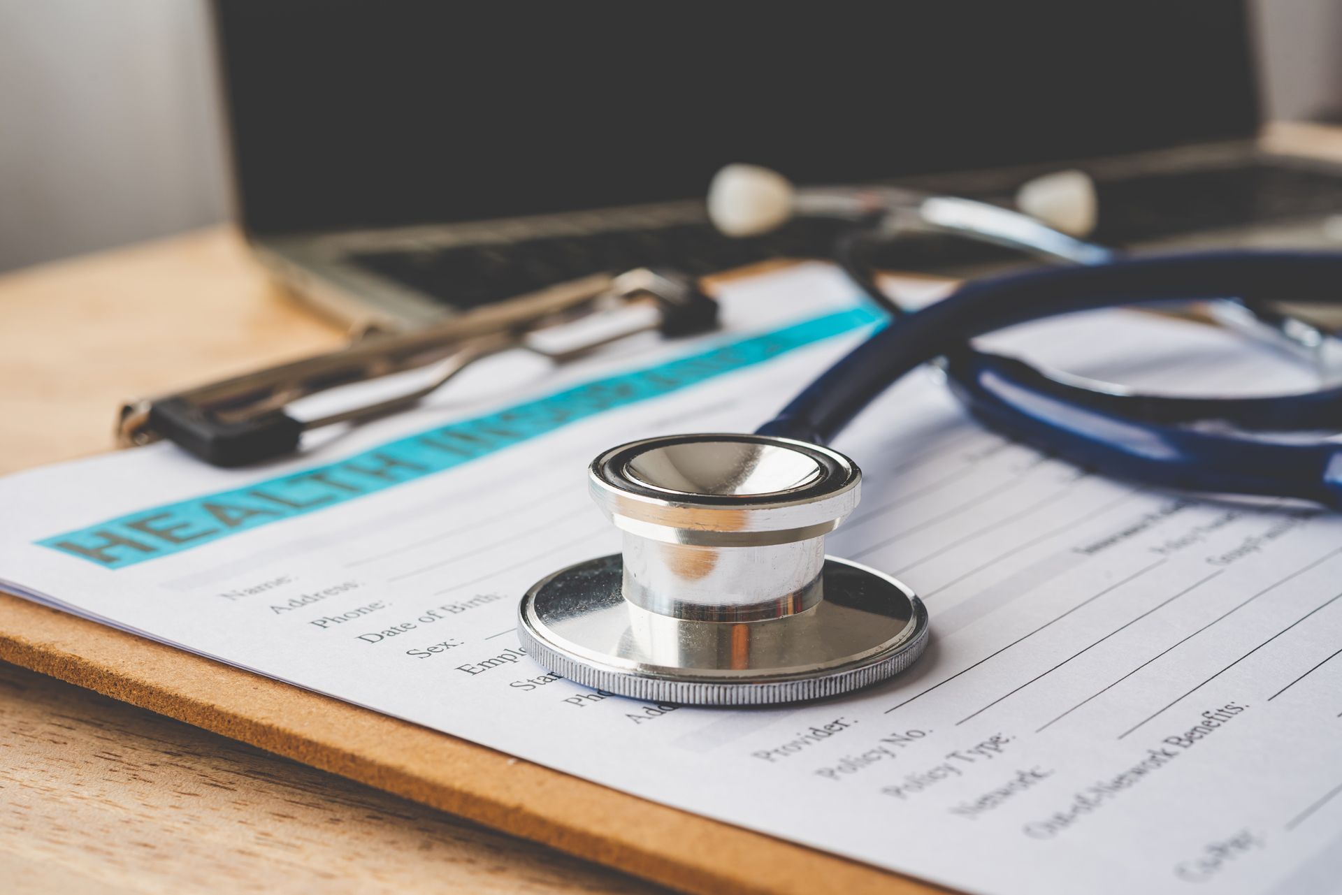 Clipboard with health form, stethoscope, glasses, and laptop on a wooden surface.