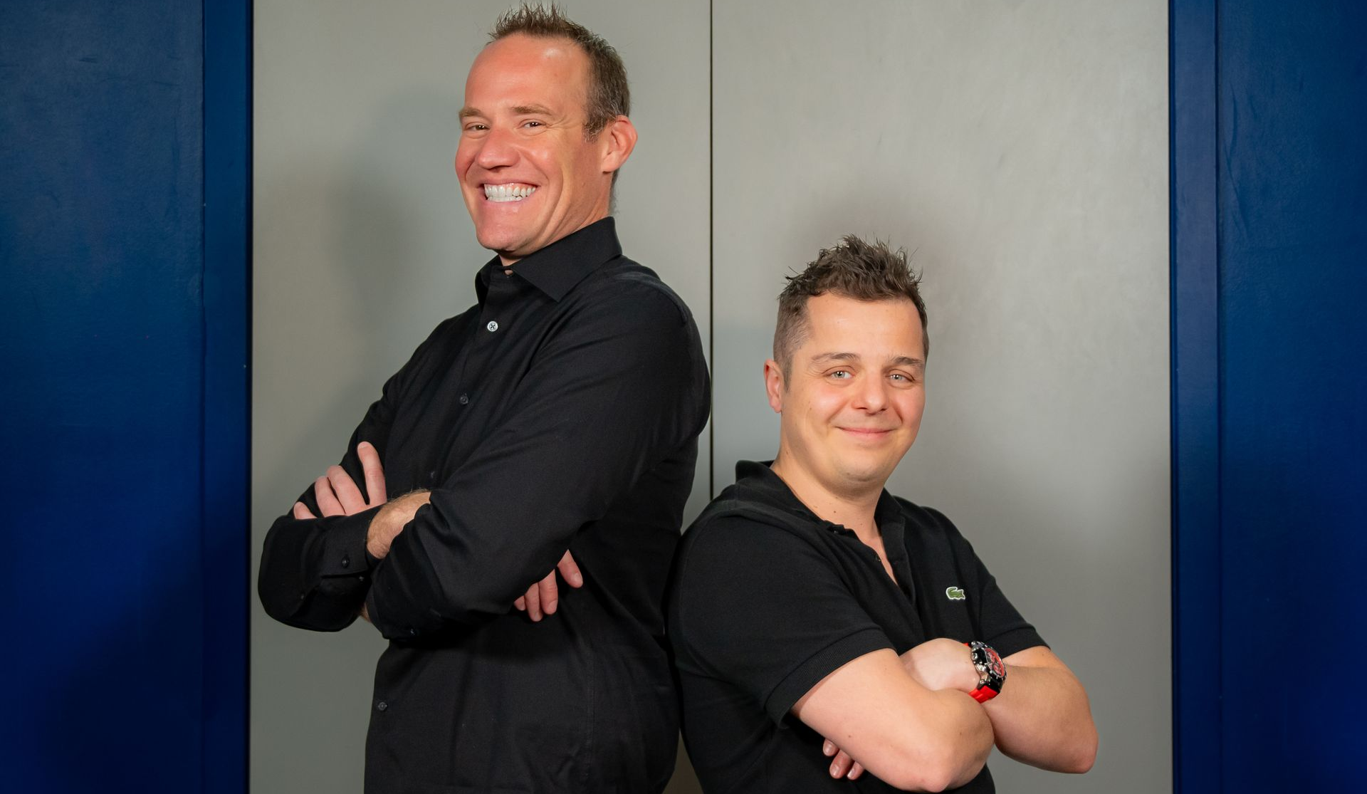 Two people in black shirts stand back-to-back against a gray wall with blue side panels, smiling at the camera.