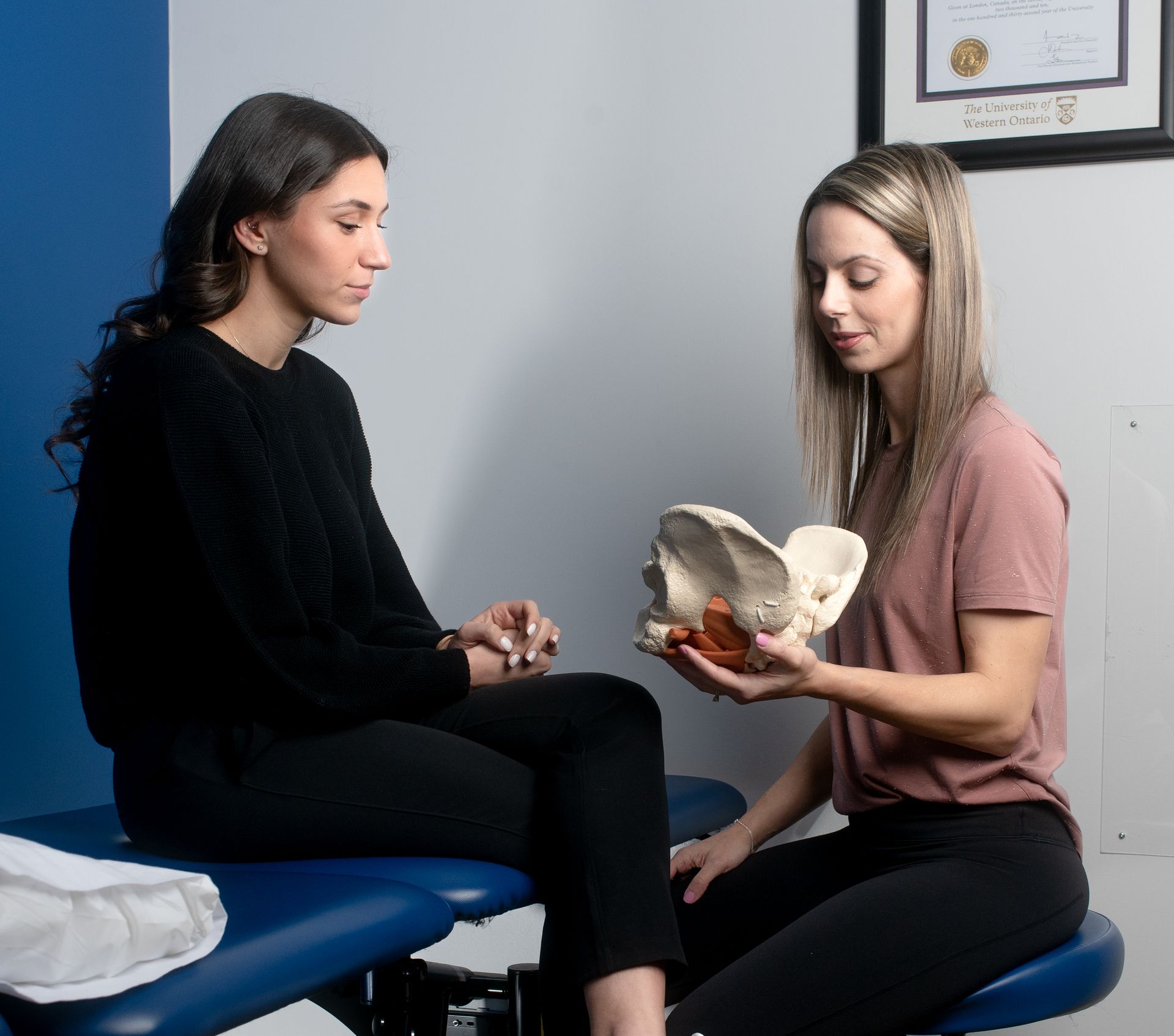A physical therapist holds a pelvic model while explaining anatomy to a patient sitting on an exam table.